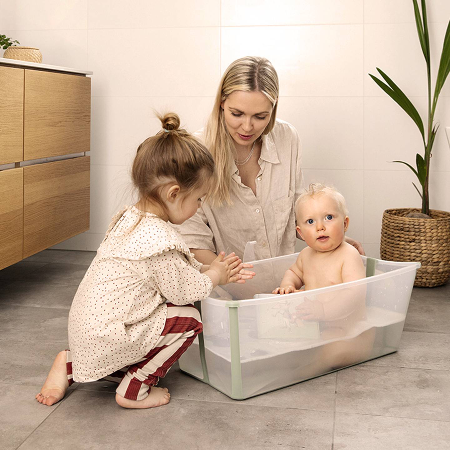 Eine Frau und ein kleines Kind baden ein Baby, das in einer Plastikwanne auf dem Badezimmerboden sitzt, neben Holzschränken und einer Topfpflanze.