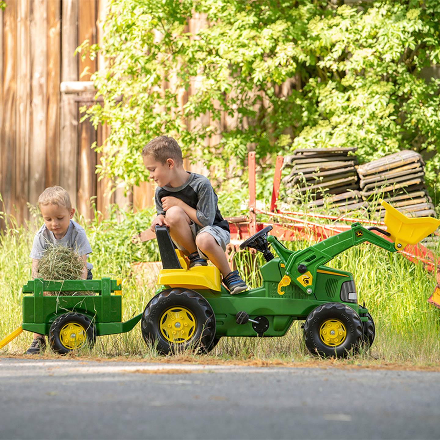 Zwei kleine Jungen spielen draußen mit einem grünen Spielzeugtraktor und Anhänger und sammeln Heu in einem sonnigen Garten mit üppigem Grün.