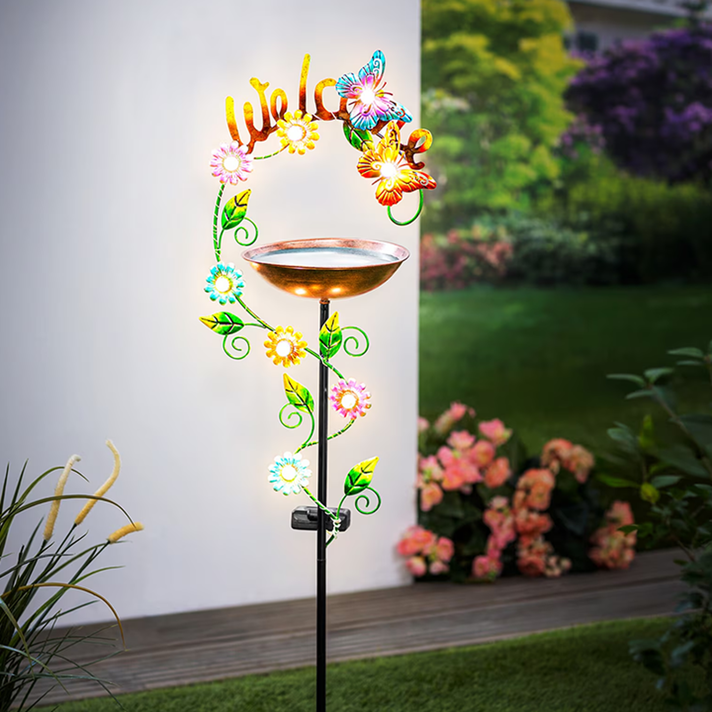 Ein dekorativer Gartenstecker mit einem Vogelbad aus Kupfer, verziert mit bunten Blumen und einem Schmetterling sowie dem Schriftzug „Welcome“, eingebettet in einen üppigen Garten.