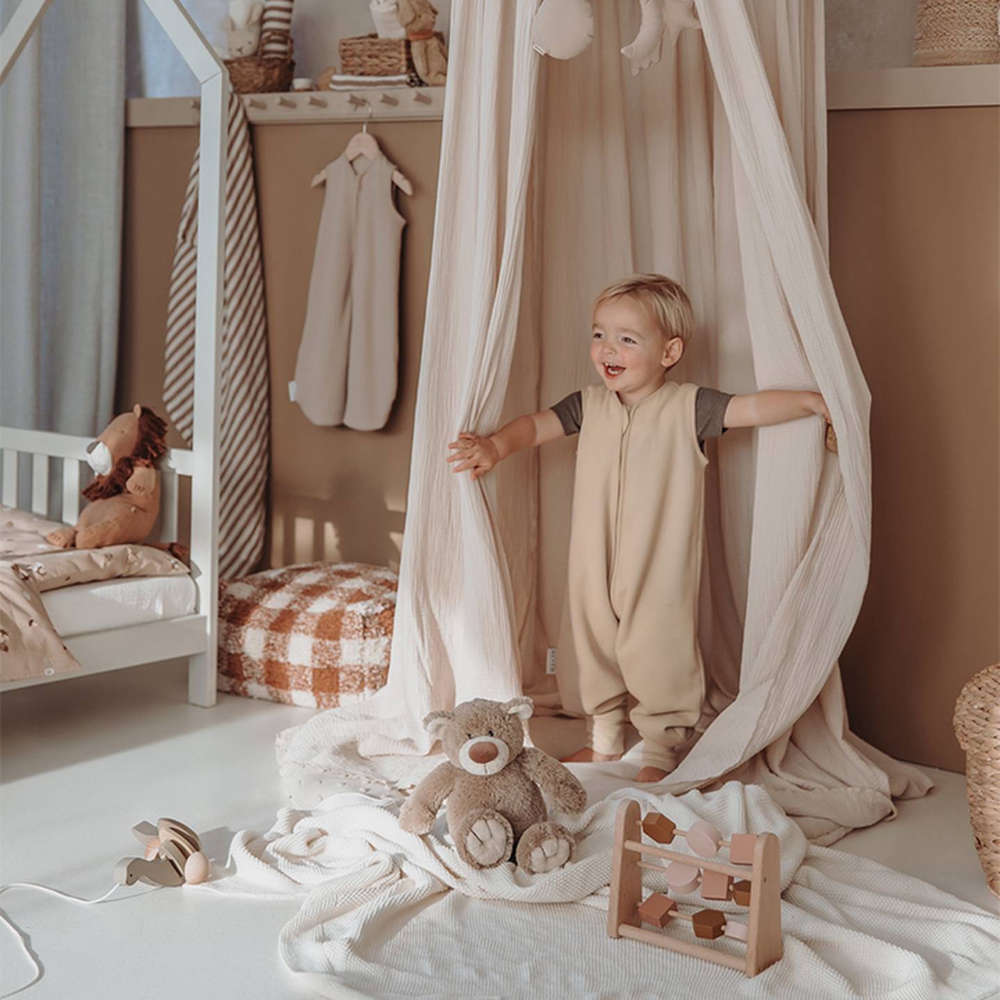 Ein Kleinkind in einem beigefarbenen Strampler spielt in einem gemütlichen, in neutralen Tönen gehaltenen Kinderzimmer mit Spielzeug, einem Baldachin und Plüschdekoration.