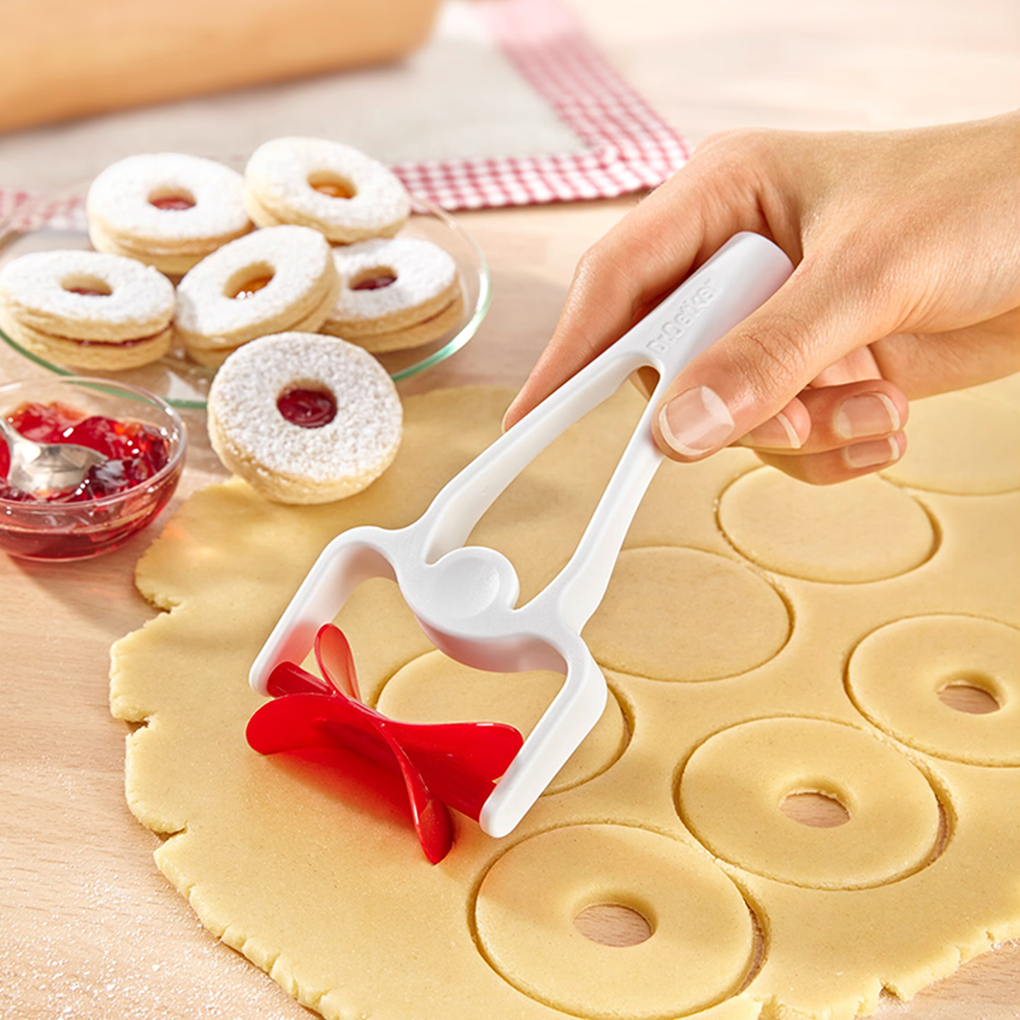 Eine Hand formt mit einem rot-weißen Teigausstecher runden Teig zu Keksen. Ein Teller mit gefüllten Keksen und eine Schale mit Marmelade stehen daneben.