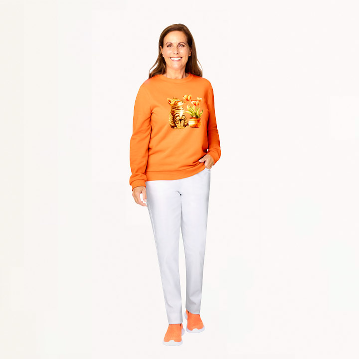 Frau in einem orangefarbenen Pullover mit Katzenmuster, weißen Hosen und orangefarbenen Schuhen, die vor einem schlichten weißen Hintergrund steht.
