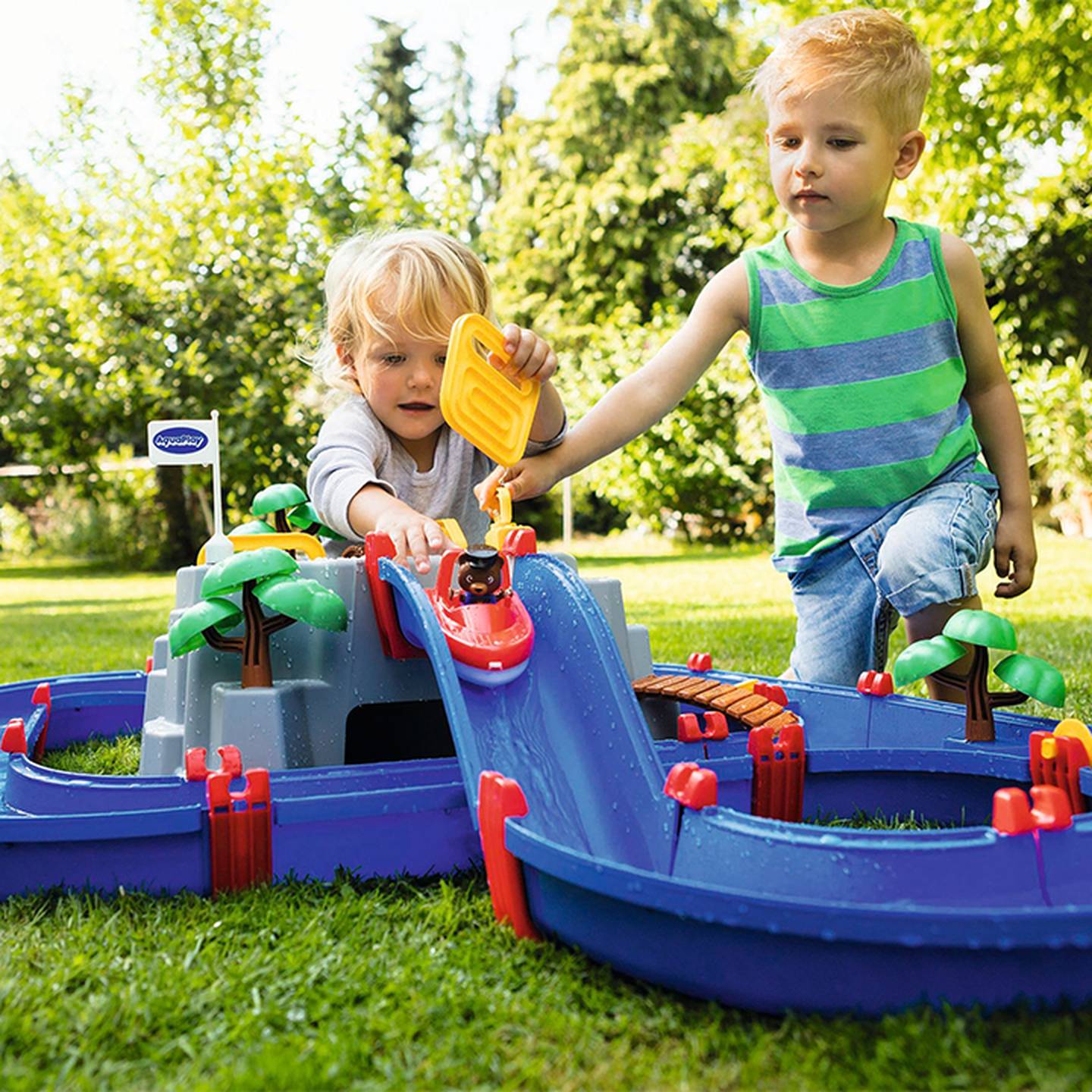 Zwei Kinder spielen im Freien auf einer Rasenfläche mit einem bunten Wasserspielset, das einen gewundenen Kanal und Spielzeugbäume zeigt.
