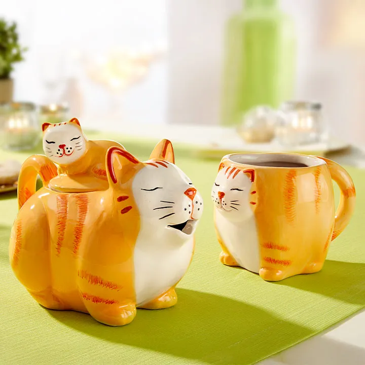 Keramik-Teekannen- und Tassenset in Form orangefarbener getigerter Katzen auf einer grünen Tischdecke, mit einer kleinen Katzenfigur auf dem Deckel der Teekanne.