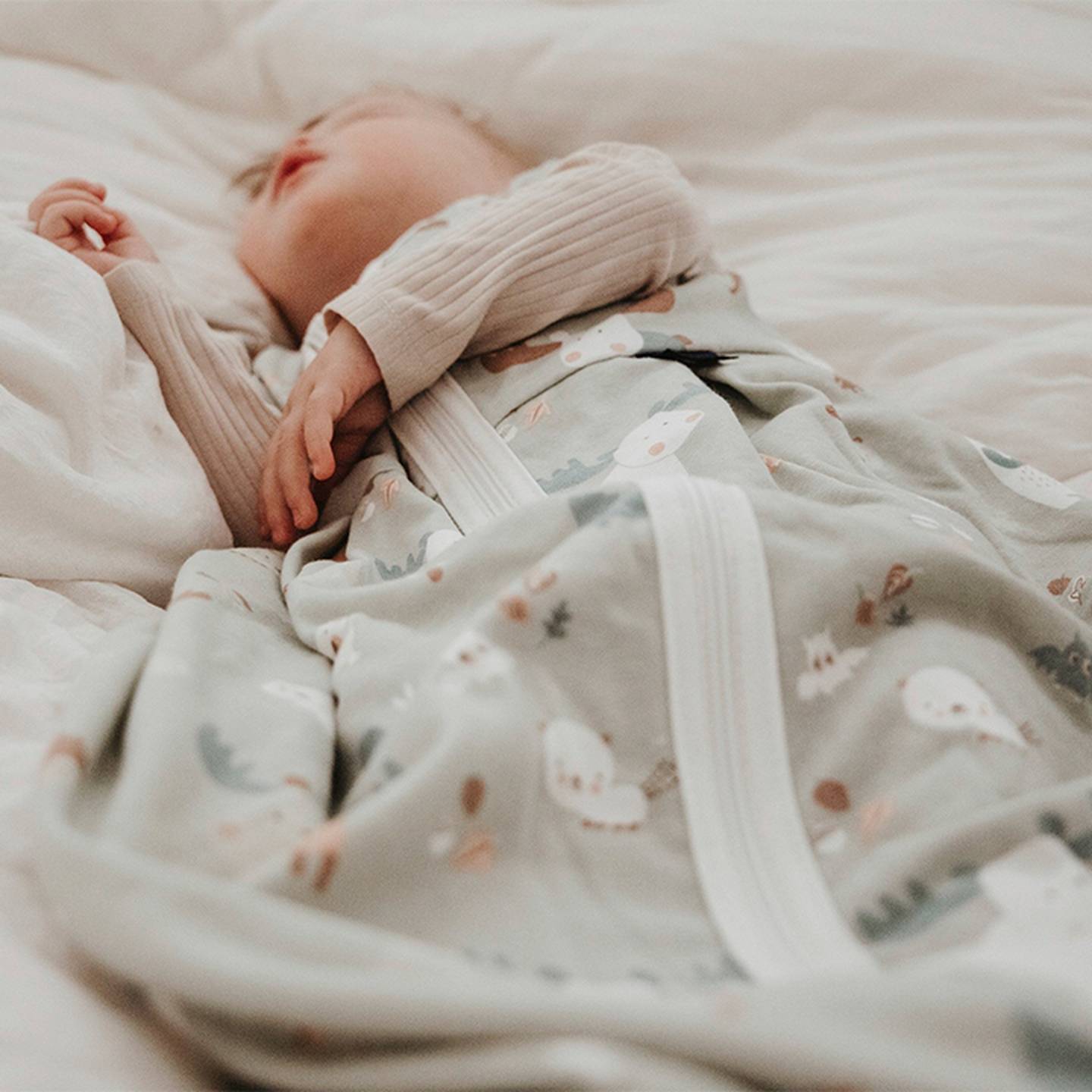 Ein Baby schläft friedlich in einem bedruckten Schlafsack auf einem weißen Bett, bekleidet mit einem gerippten, beigefarbenen Langarmshirt.