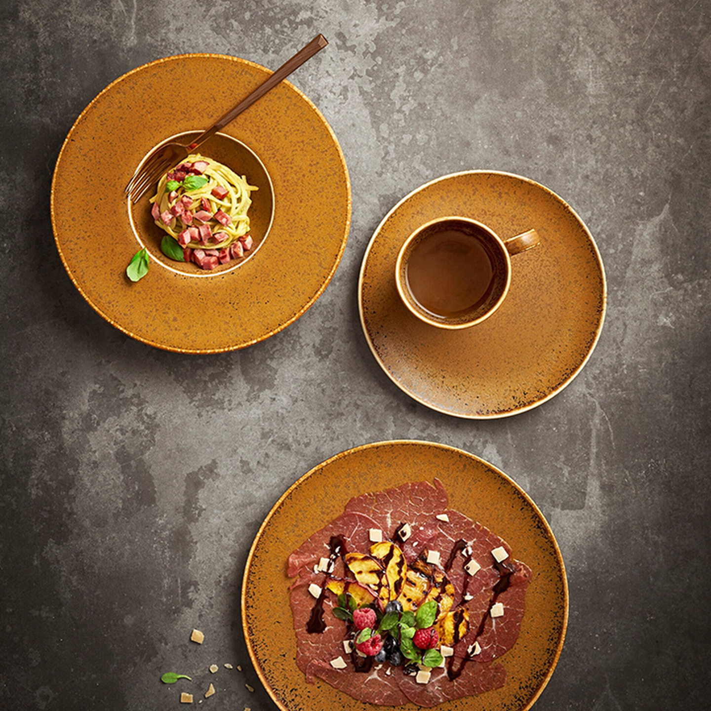 Luftaufnahme von drei rustikalen Tellern auf einer Schieferplatte: eine Tasse Kaffee, Spaghetti mit Garnitur sowie ein Gericht mit Fleischscheiben, Obst und einer Sauce.