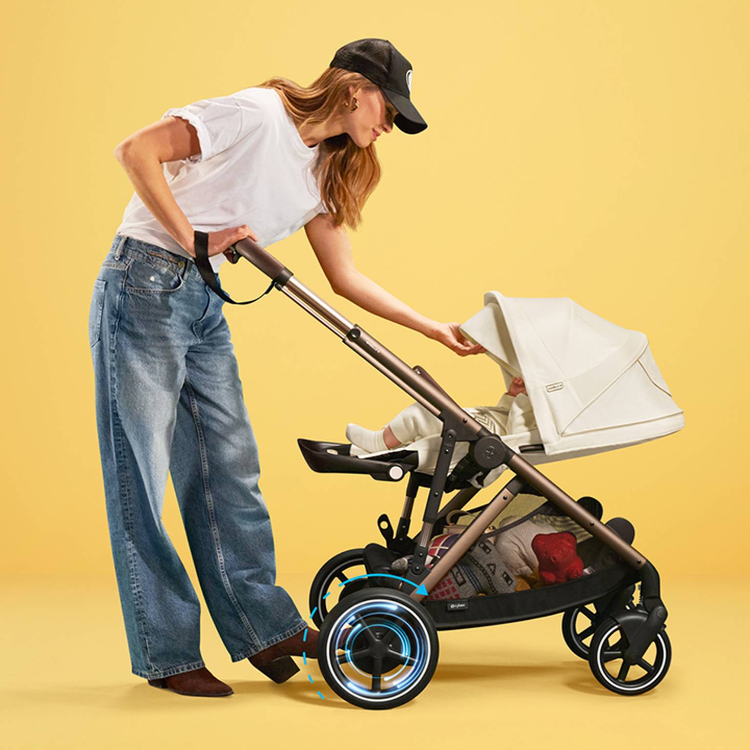 Frau in weißem Hemd und Jeans stellt einen Kinderwagen mit einem Baby darin vor einem gelben Hintergrund ein. Der Kinderwagen verfügt über hervorgehobene Räder.