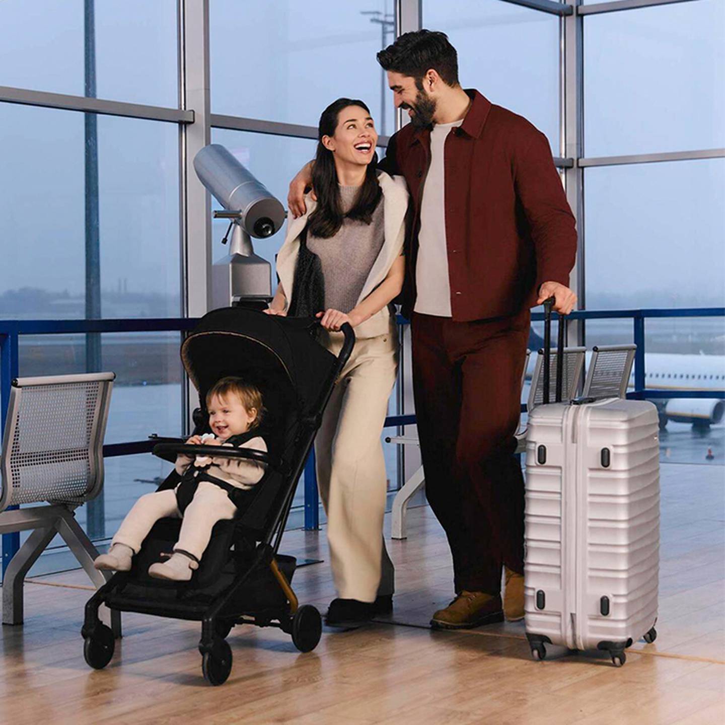 Eine Familie am Flughafen: lächelnde Eltern mit einem Kleinkind im Kinderwagen, Gepäck in der Nähe und ein großes Fenster mit Blick auf die Start- und Landebahn.