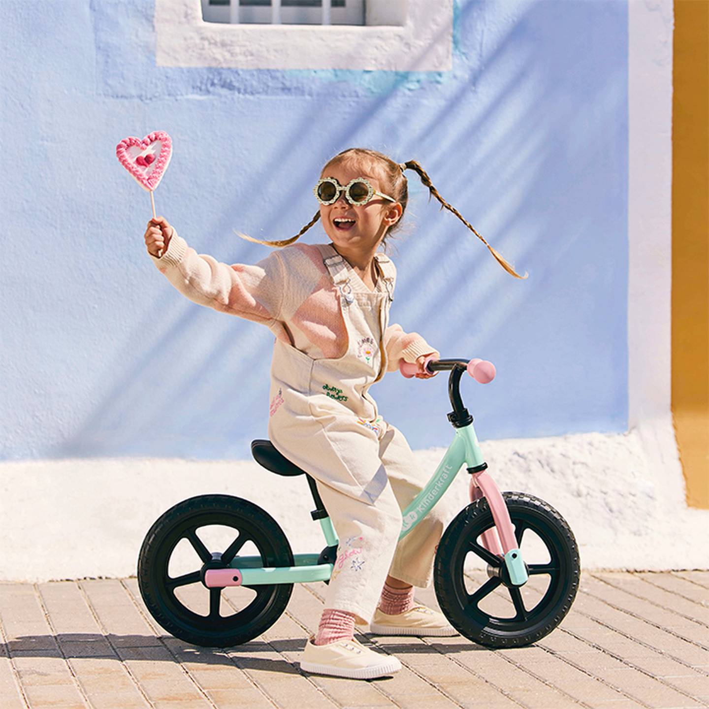 Ein fröhliches Kind mit Zöpfen fährt auf einem Laufrad, schwenkt dabei einen rosa Herzstab, trägt eine Sonnenbrille und ein pastellfarbenes Outfit und steht vor einer blau-gelben Wand.