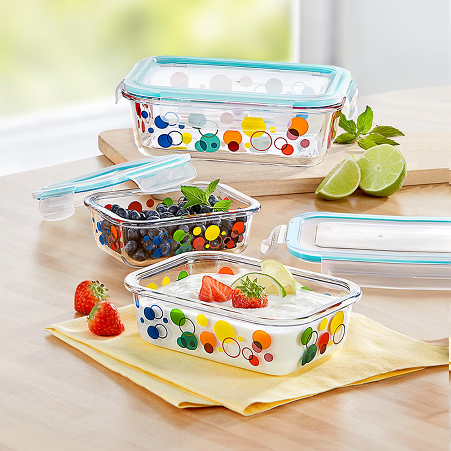 Bunte Glasbehälter mit Joghurt, Obst und Beeren – garniert mit Minze und Limette – auf einer Holzoberfläche mit lebhaften gepunkteten Mustern.