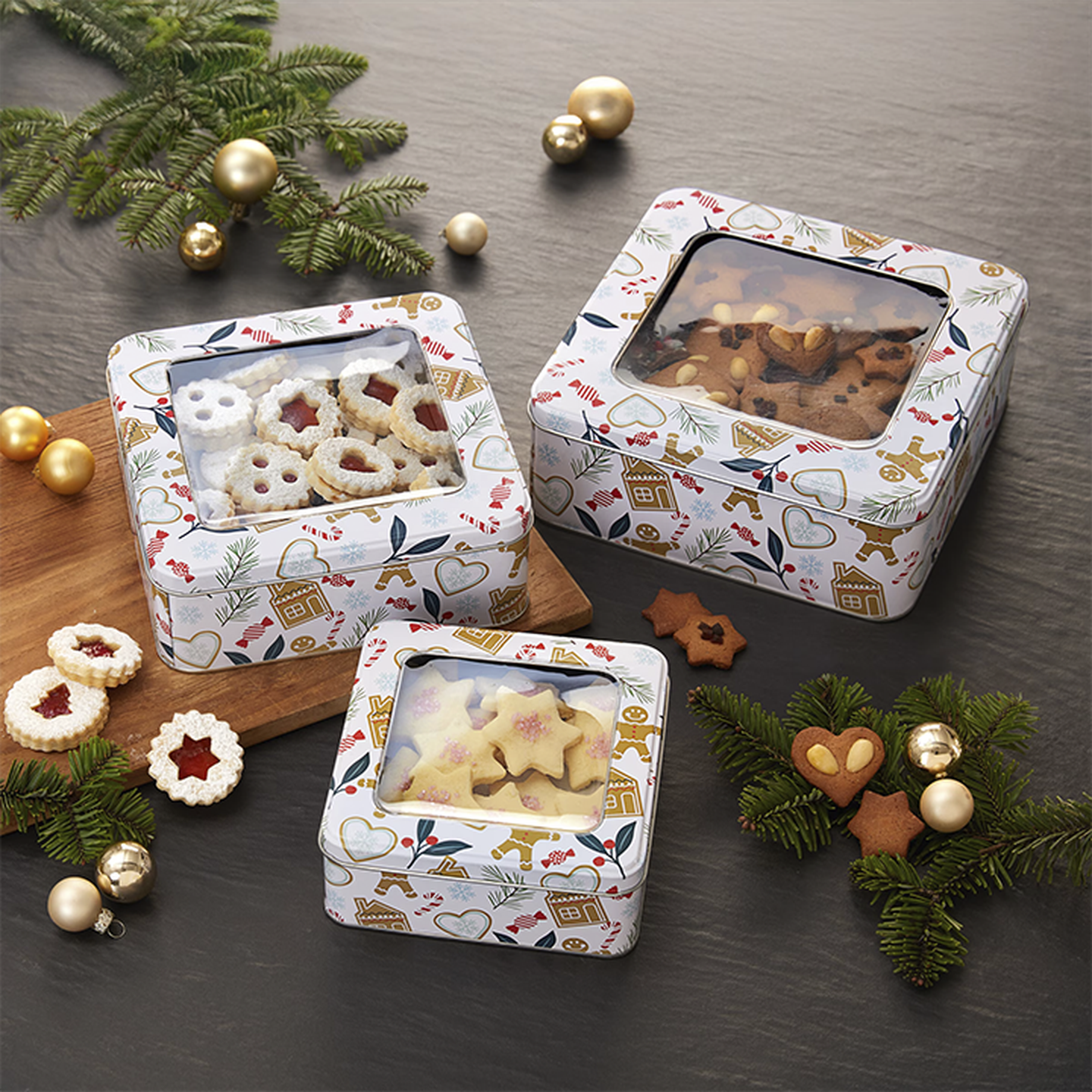 Drei festliche Dosen mit verschiedenen Keksen, umgeben von Tannenzweigen und goldenen Ornamenten auf dunklem Untergrund.