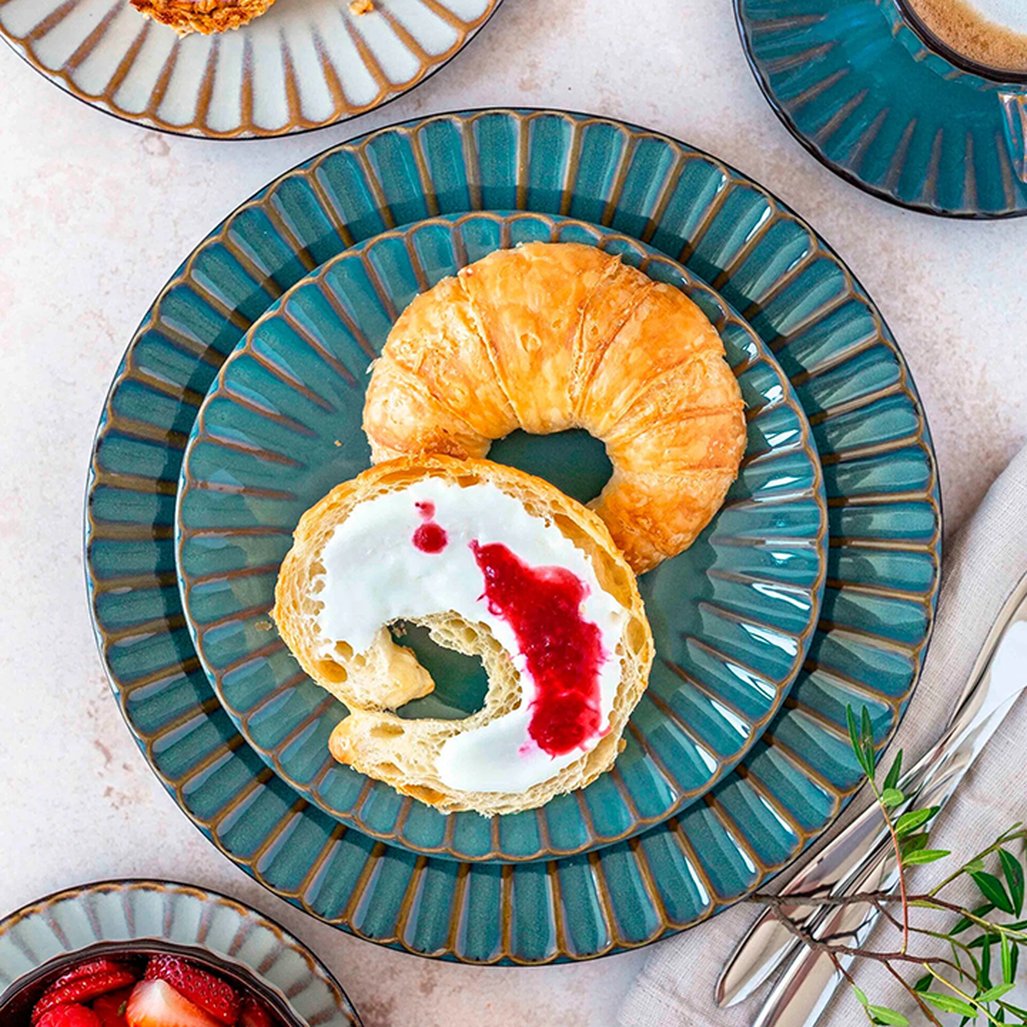 Ein Croissant mit Sahne und Himbeersauce auf einem blauen Teller, umgeben von Kaffee, Besteck und einer Beilage aus Erdbeerscheiben.