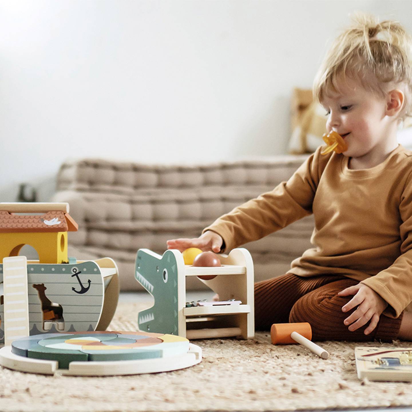Ein Kleinkind mit Schnuller spielt in einem gemütlichen Wohnzimmer auf dem Boden mit bunten Holzspielzeugen, darunter ein Krokodil und ein Puzzle.
