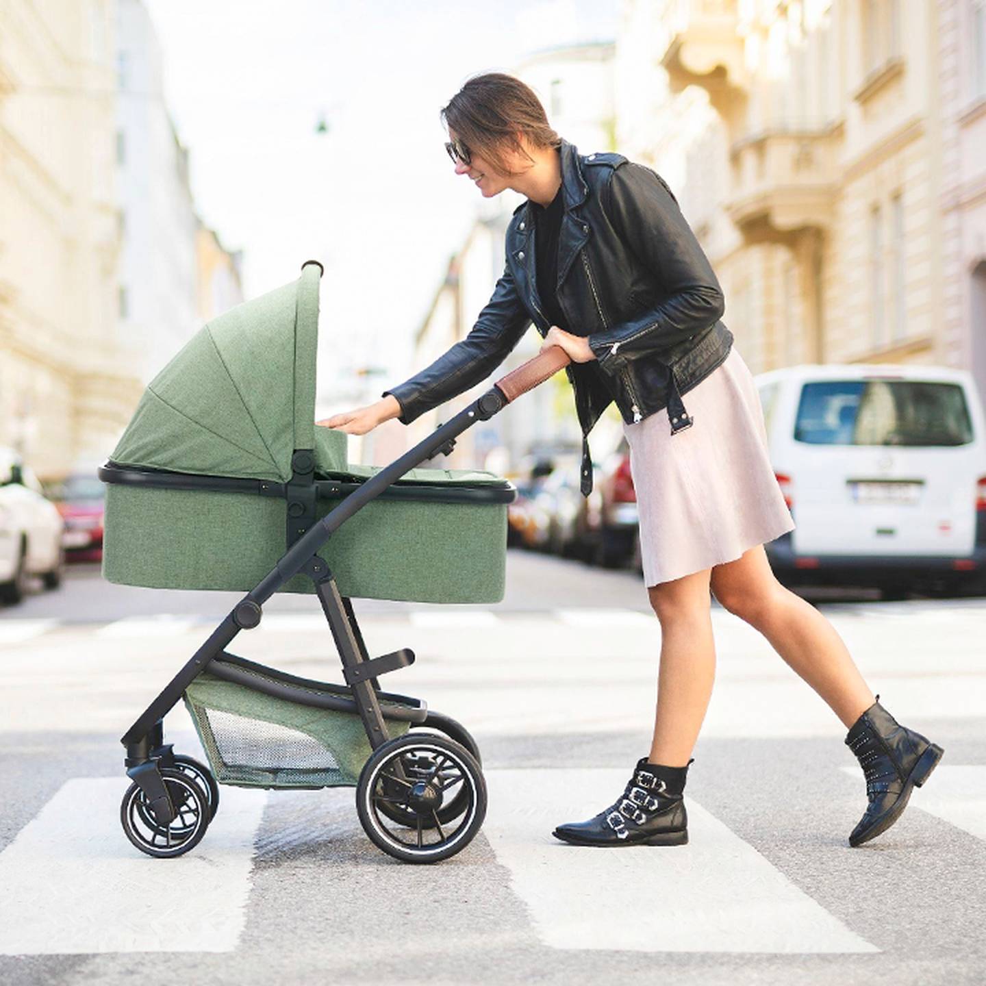 Eine Frau in einer Lederjacke schiebt einen grünen Kinderwagen über eine Stadtstraße, umgeben von geparkten Autos und Gebäuden.