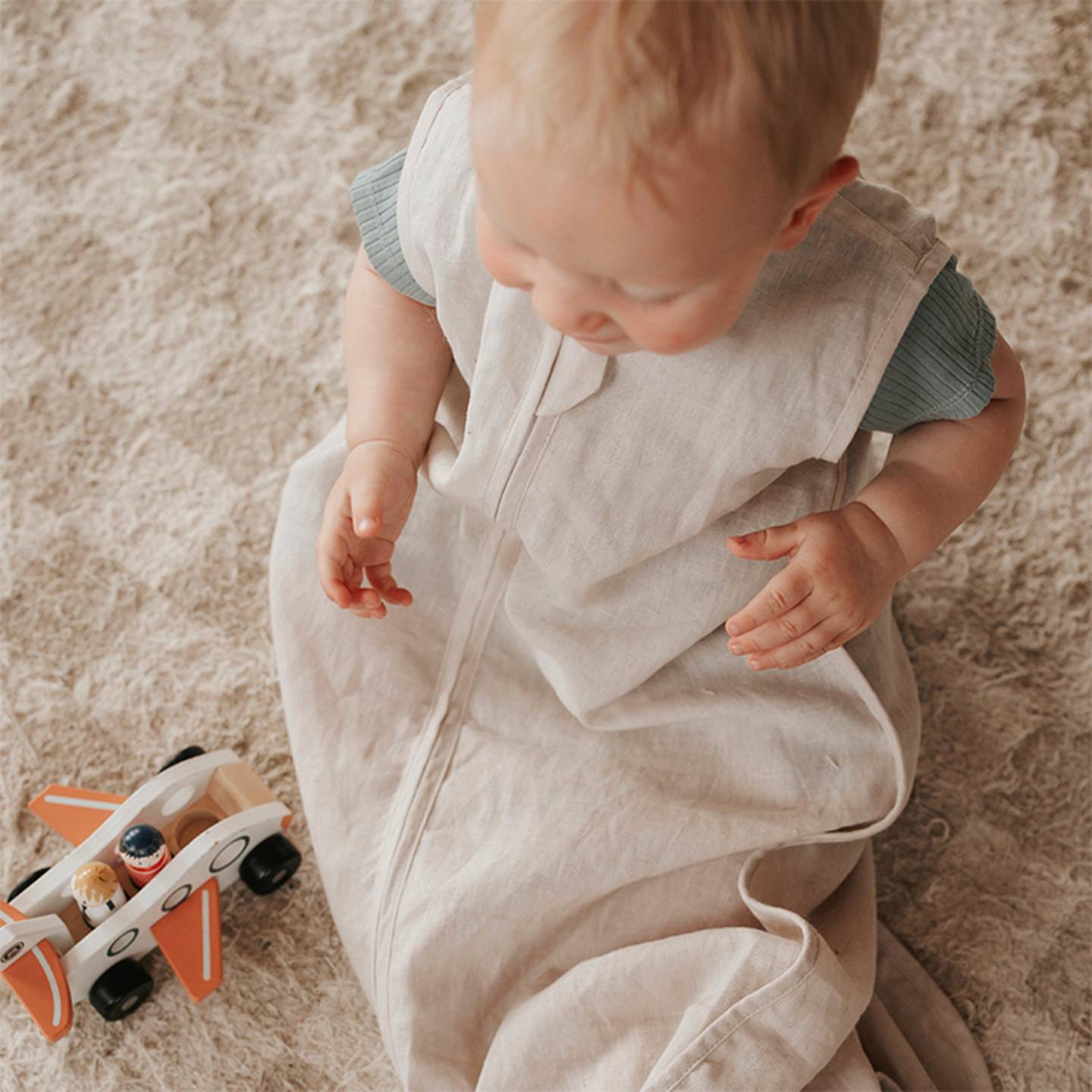 Ein Baby in einem hellbeigen Schlafsack sitzt auf einem strukturierten Teppich neben einem Holzspielzeugflugzeug mit orangefarbenen Akzenten.