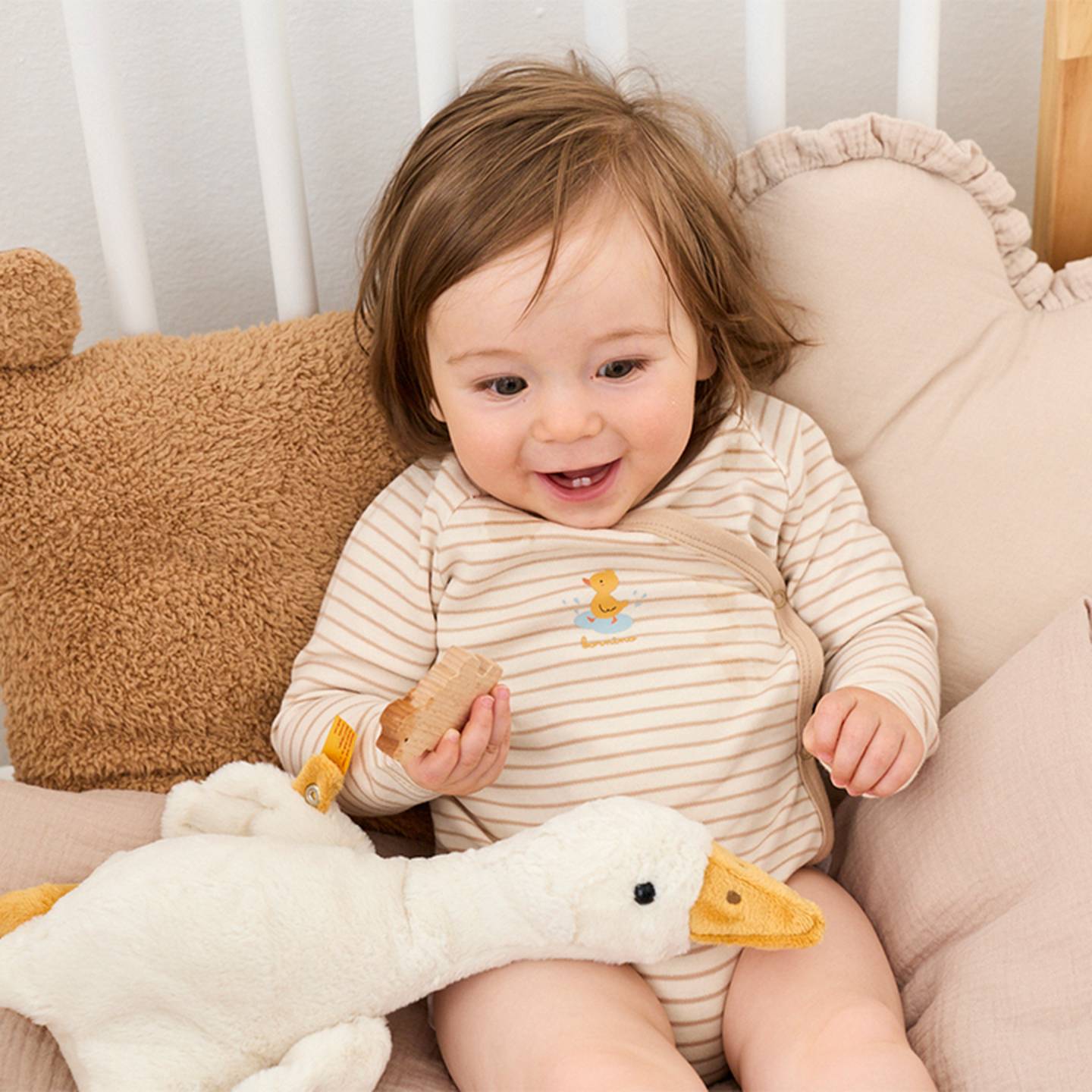 Baby sitzt neben einer Kuscheltier Ente im Bett.