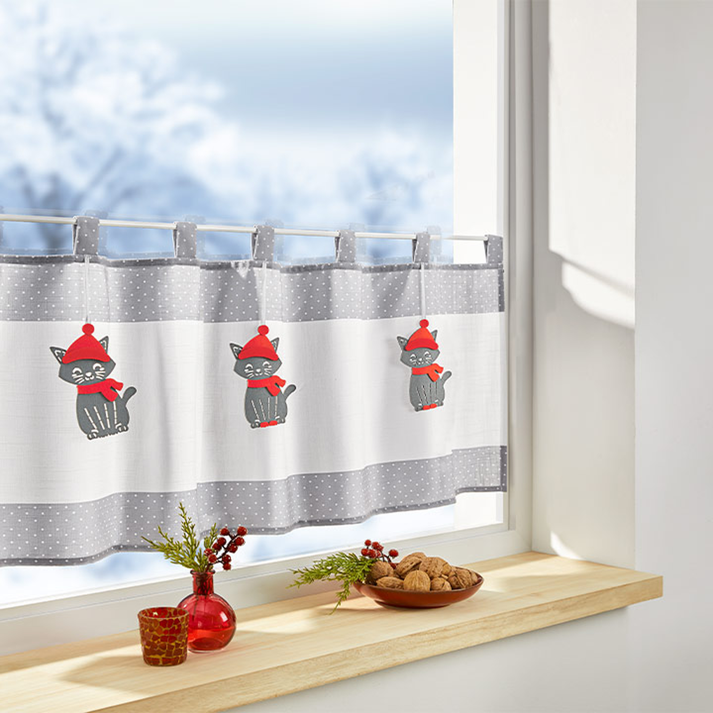 Weiß-grauer Küchenvorhang mit niedlichen Katzenmotiven und roten Akzenten. Auf einer Fensterbank stehen eine rote Vase mit Grünpflanzen und eine Schale mit Walnüssen.