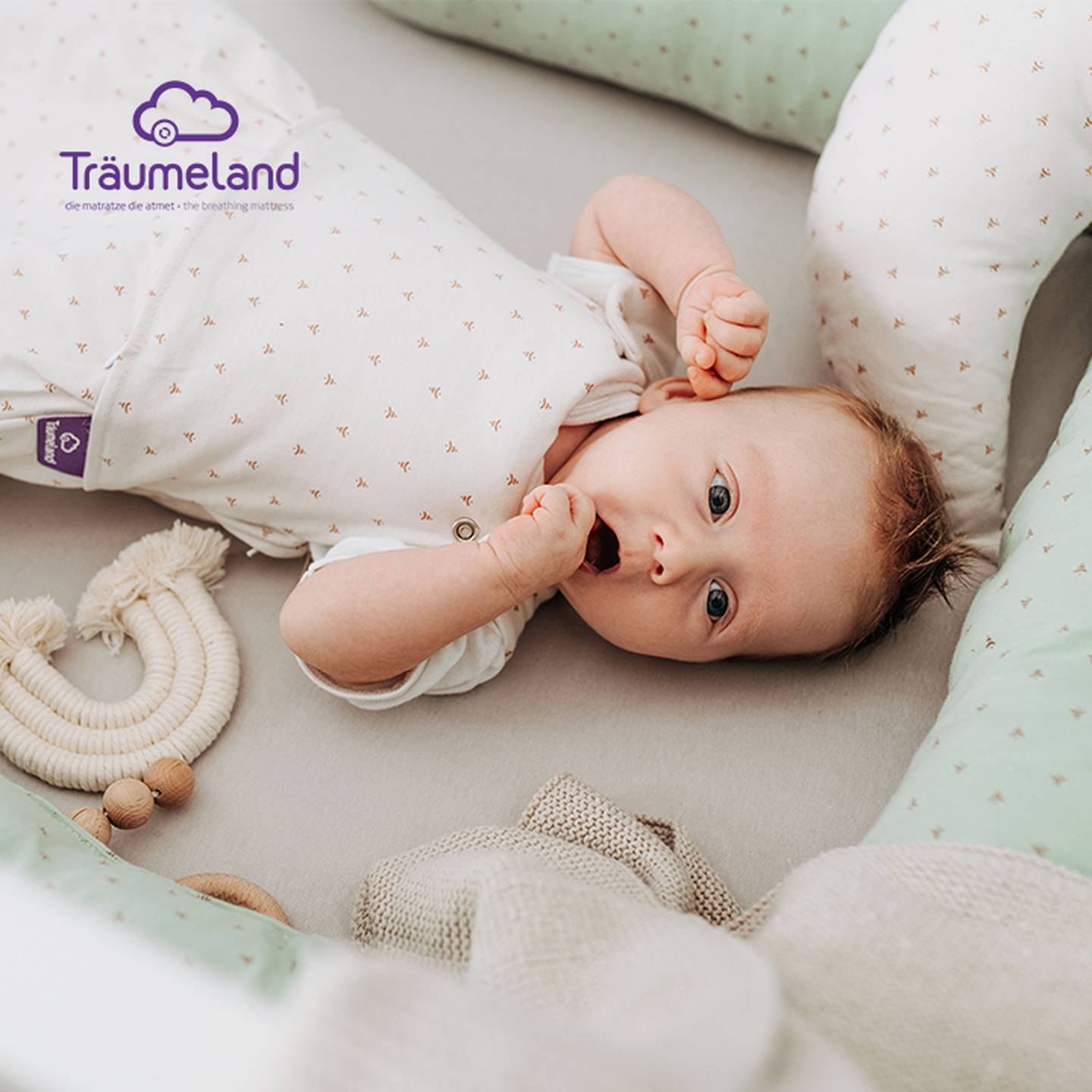 Baby liegt in einem Kinderbett, trägt einen weißen Strampler und ist von weichen Kissen und Spielzeug umgeben. In der oberen linken Ecke ist das Logo „Träumeland“ zu sehen.