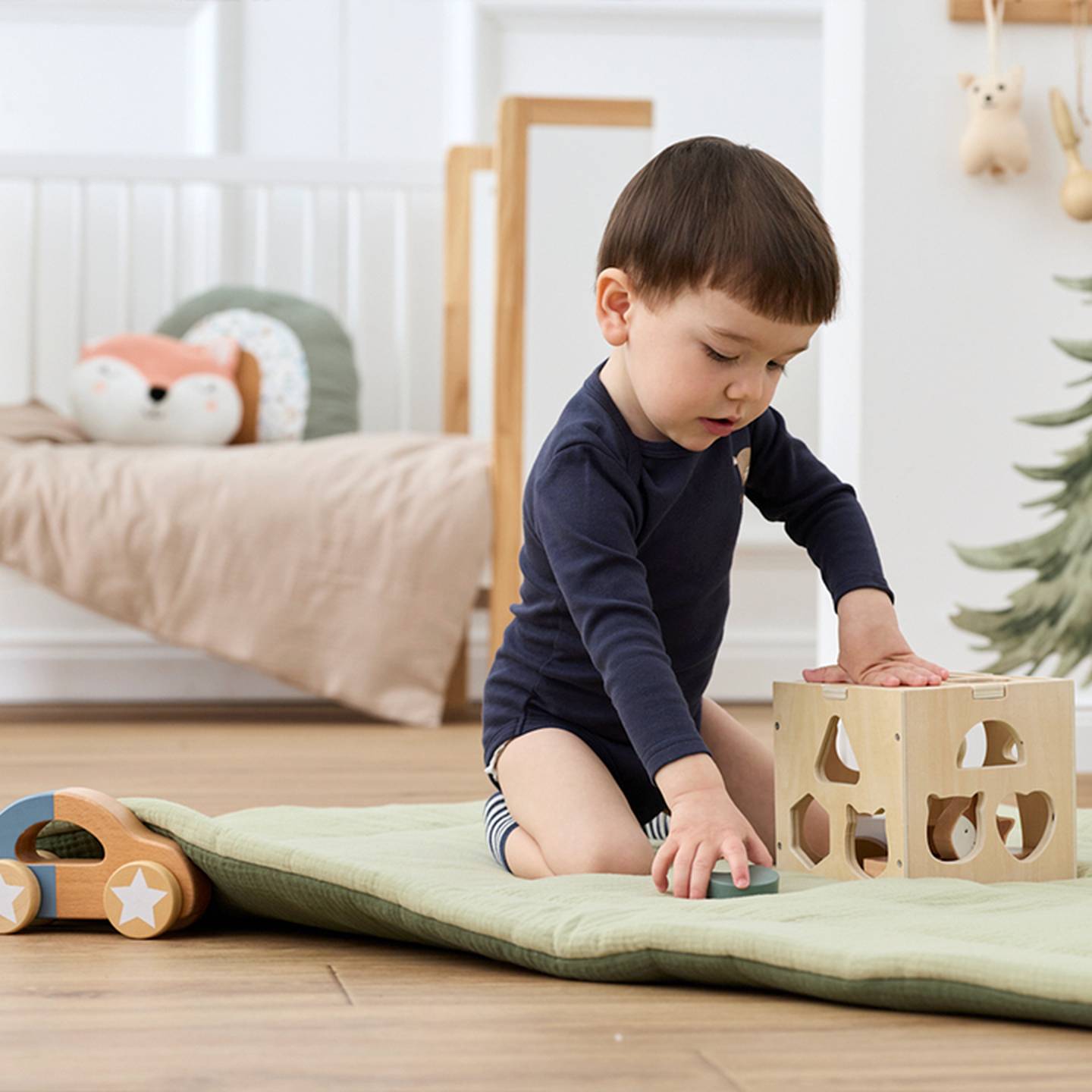 Ein Kleinkind in einem marineblauen Strampler spielt auf einer gepolsterten Matte mit einem hölzernen Formensortierer. In der Nähe steht ein hölzernes Spielzeugauto, und im Hintergrund ist ein gemütliches Bett zu sehen.