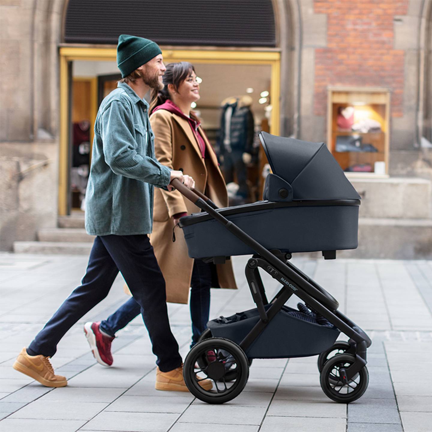 Ein Paar geht lächelnd eine Stadtstraße entlang. Der Mann schiebt einen dunklen Kinderwagen. Sie tragen legere, warme Kleidung.
