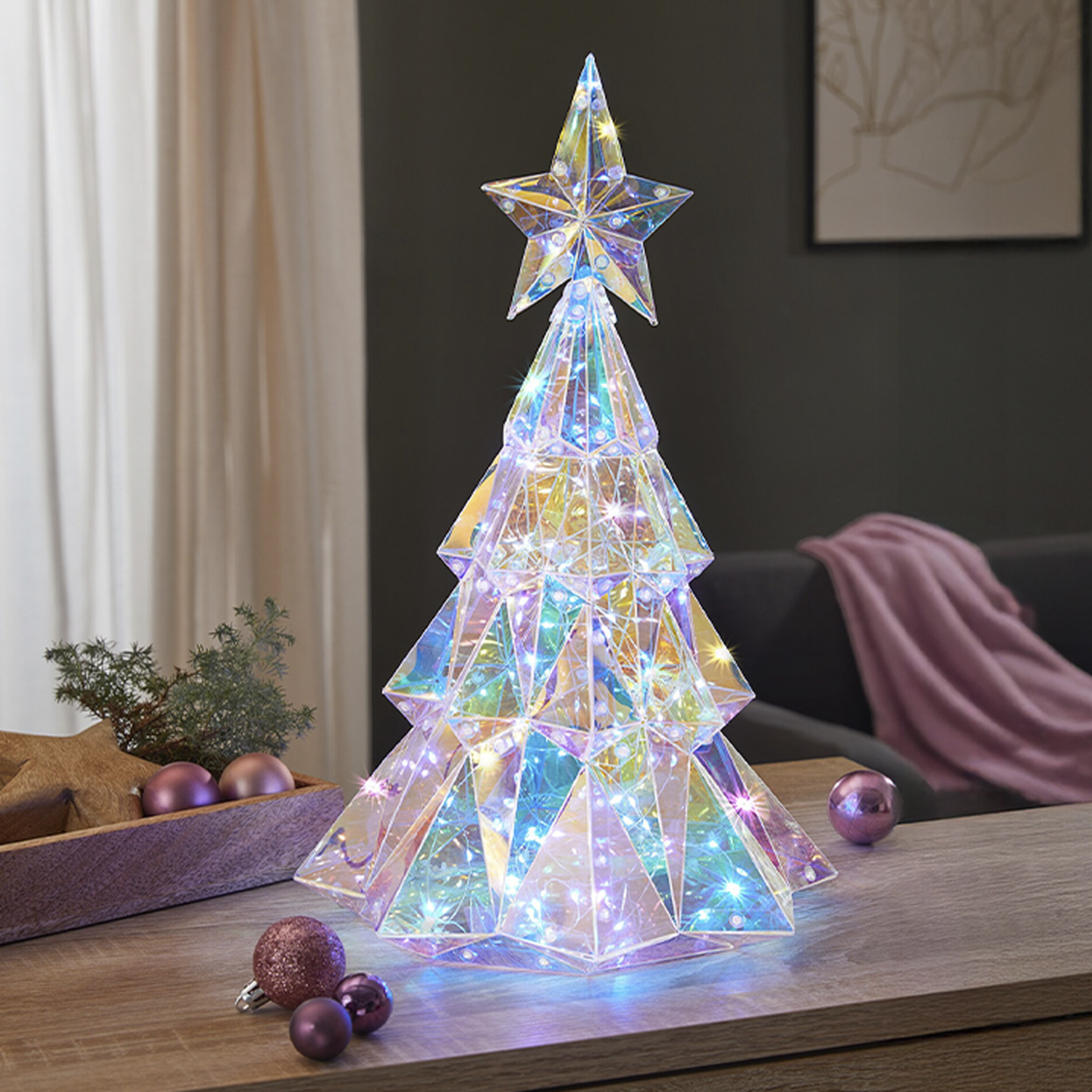 Ein leuchtender, mehrfarbiger LED-Weihnachtsbaum mit einer Sternspitze auf einem Holztisch, umgeben von rosa Ornamenten und einer kleinen Pflanze.