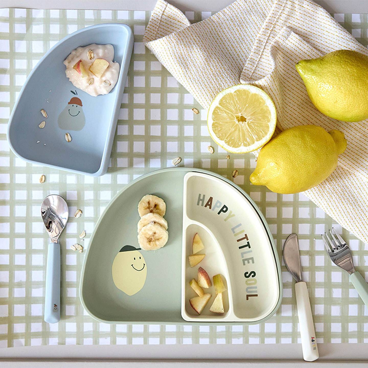 Kinderteller mit fröhlichen Gesichtern, geschnittene Bananen und Äpfel, umgeben von Zitronen und Besteck auf einer karierten Tischdecke.