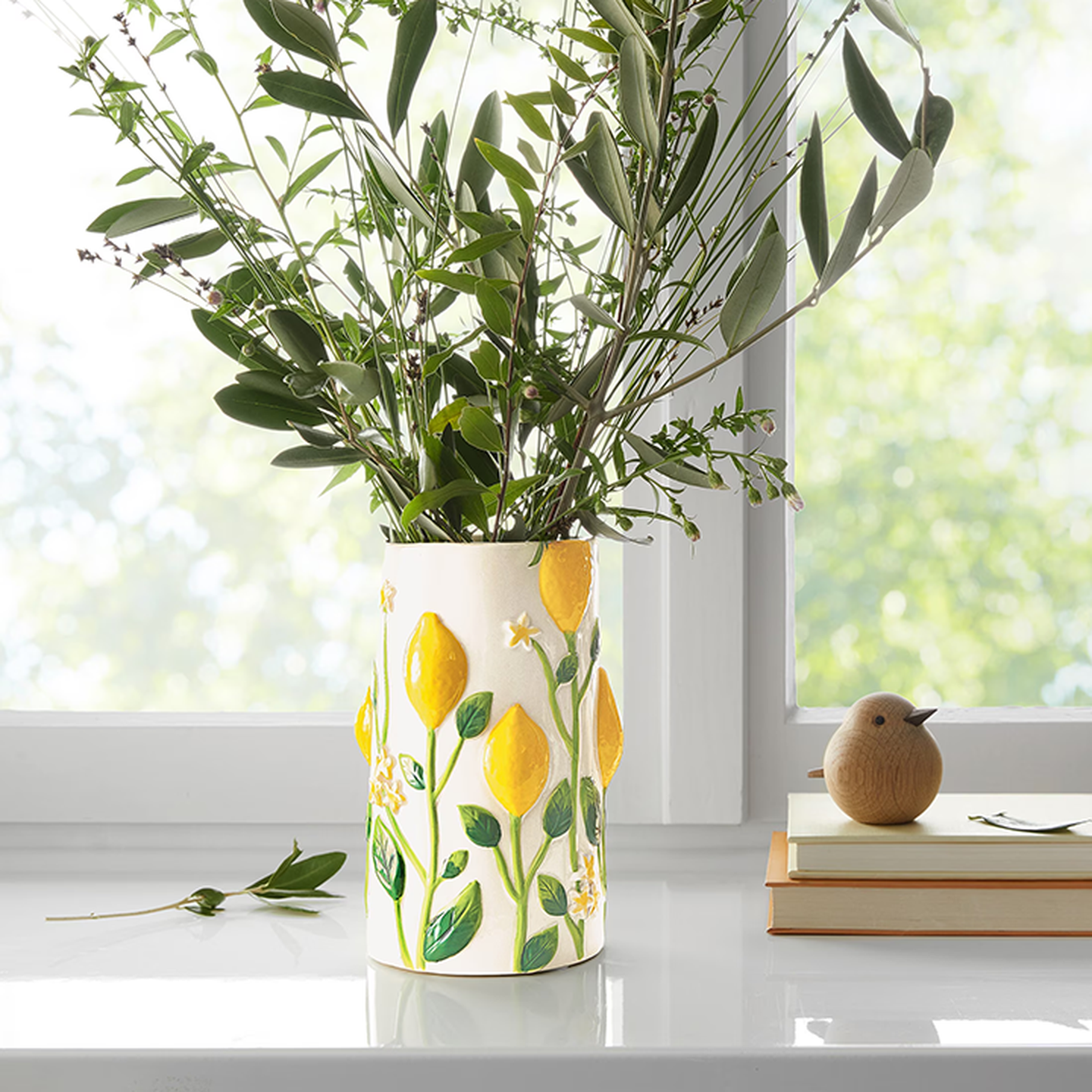 Eine dekorative Vase mit gelben Zitronen und grünen Blättern enthält frisches Grün und steht auf einer Fensterbank, neben einem Bücherstapel und einer hölzernen Vogelfigur.