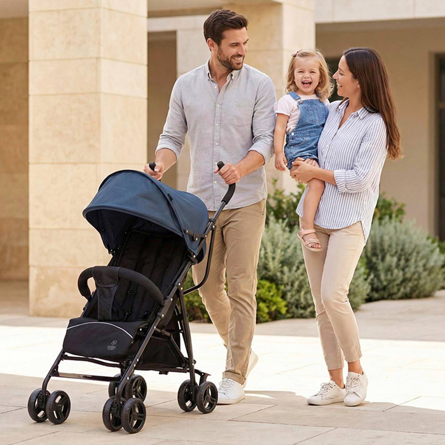 Eine lächelnde Familie spaziert im Freien; der Vater schiebt einen Kinderwagen, die Mutter hält ein lachendes Kleinkind im Arm. Sie tragen legere Kleidung.