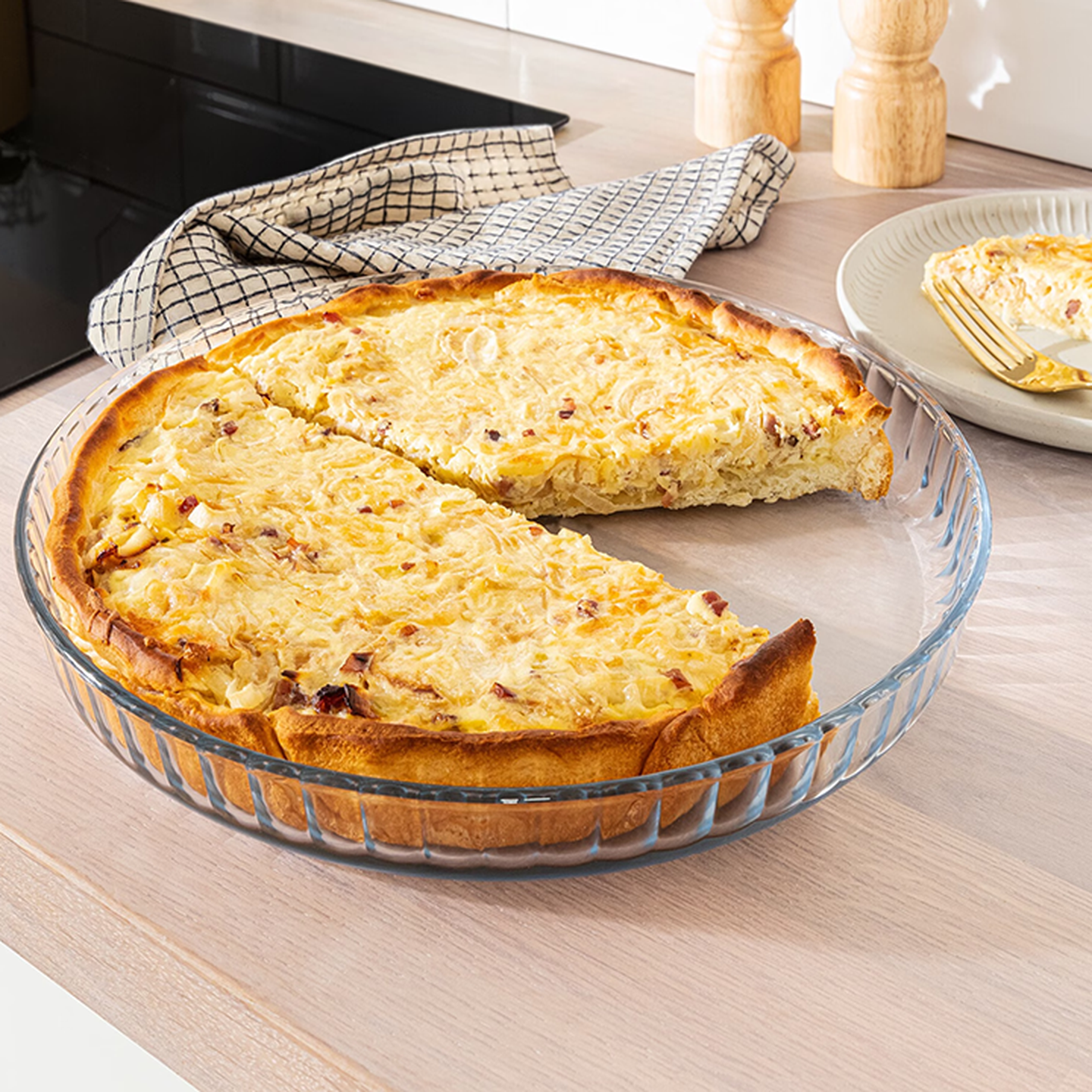 Eine Käse-Speck-Quiche, bei der ein Stück fehlt; sie steht auf einer Holztheke neben einem Teller mit Serviette und Gabel.