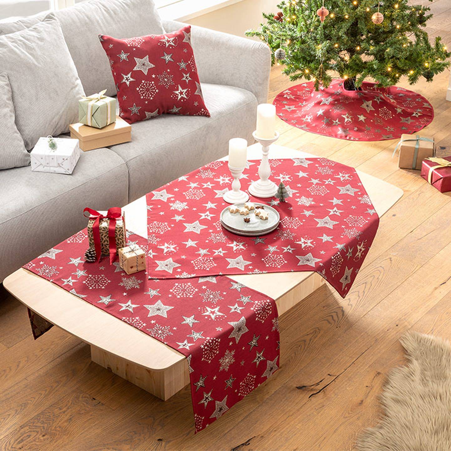 Festlich eingerichtetes Wohnzimmer mit rotem, sternenmusterbesetztem Tisch und Baumdecken, Geschenken, Kerzen und einem geschmückten Weihnachtsbaum neben einem grauen Sofa.