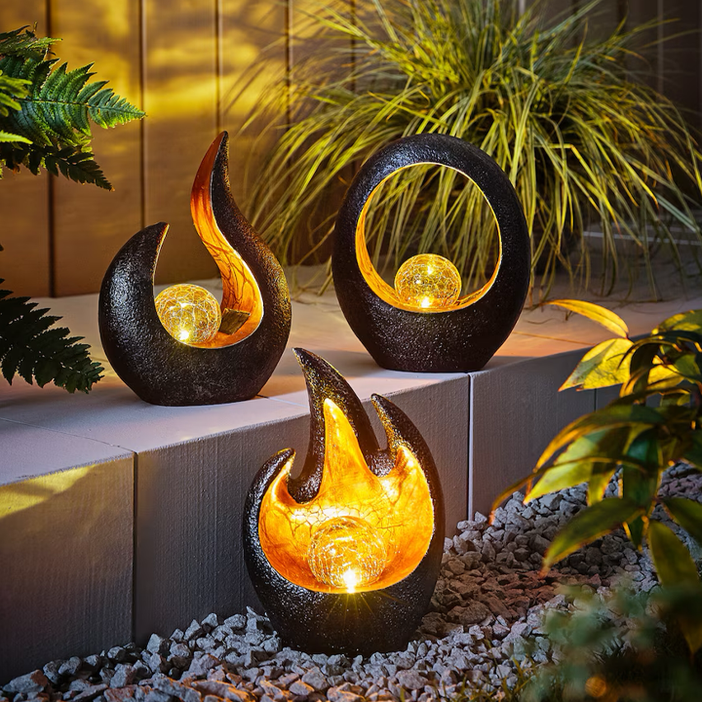 Drei dekorative Gartenleuchten auf einem Steinweg, jede mit einer leuchtenden Kugel, die von schwarzen und goldenen Skulpturen umgeben ist und von Laubwerk umrahmt wird.