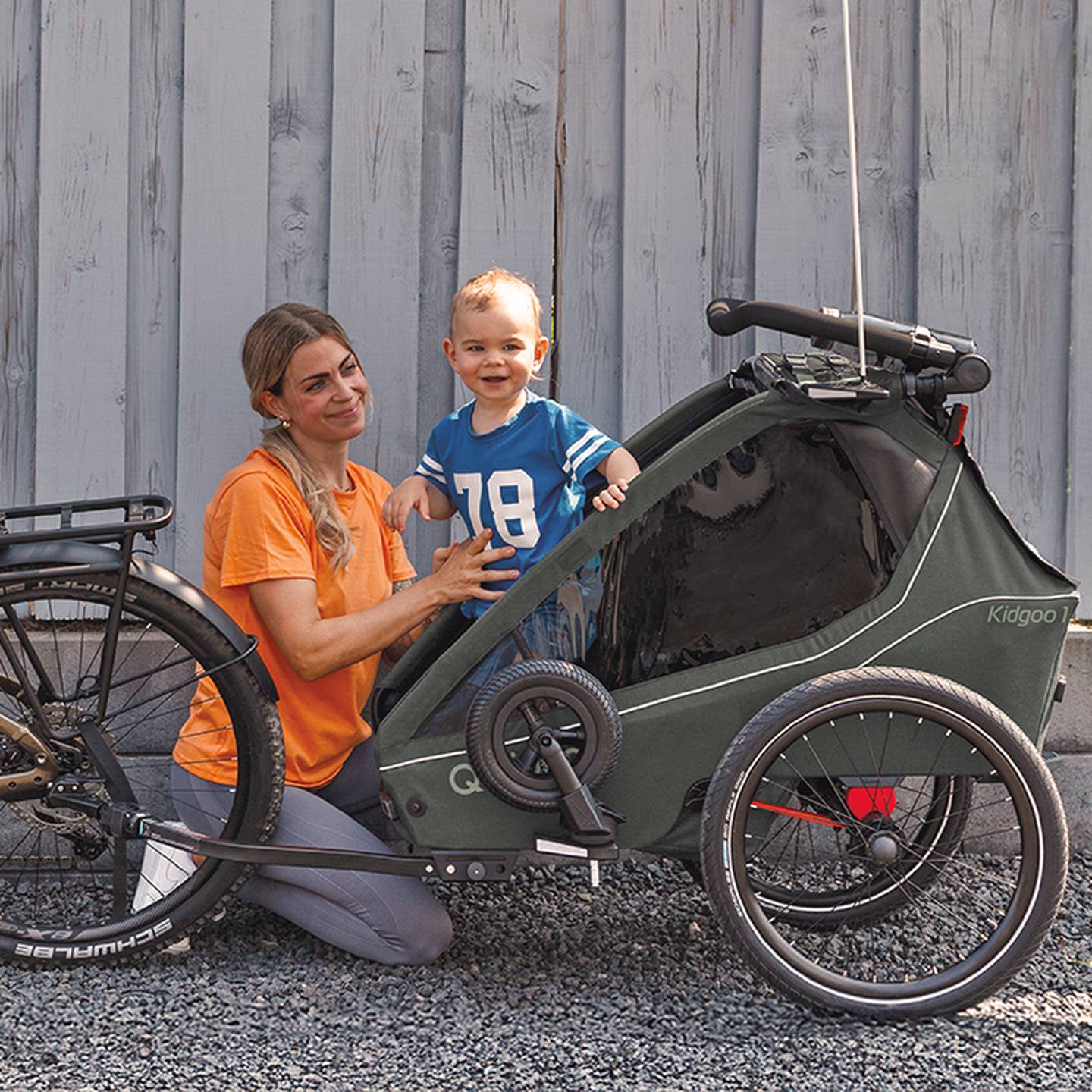 Eine Frau kniet neben einem Kleinkind in einem Fahrradanhänger. Das Kleinkind trägt ein blaues Hemd und lächelt. Der Anhänger ist an einem Fahrrad befestigt.