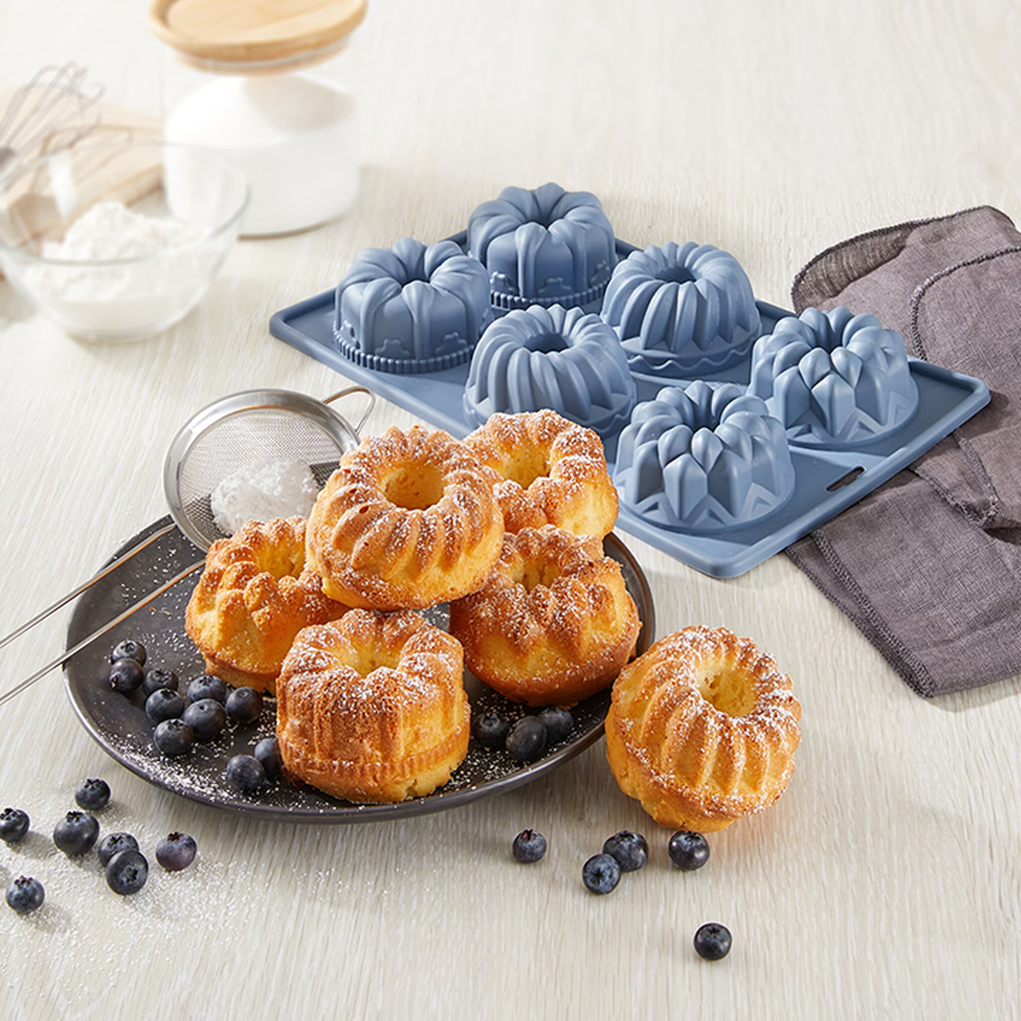 Mit Puderzucker bestäubte Mini-Gugelhupfe auf einem Teller, umgeben von Blaubeeren, neben einer blauen Silikonform auf einem hellen Holztisch.
