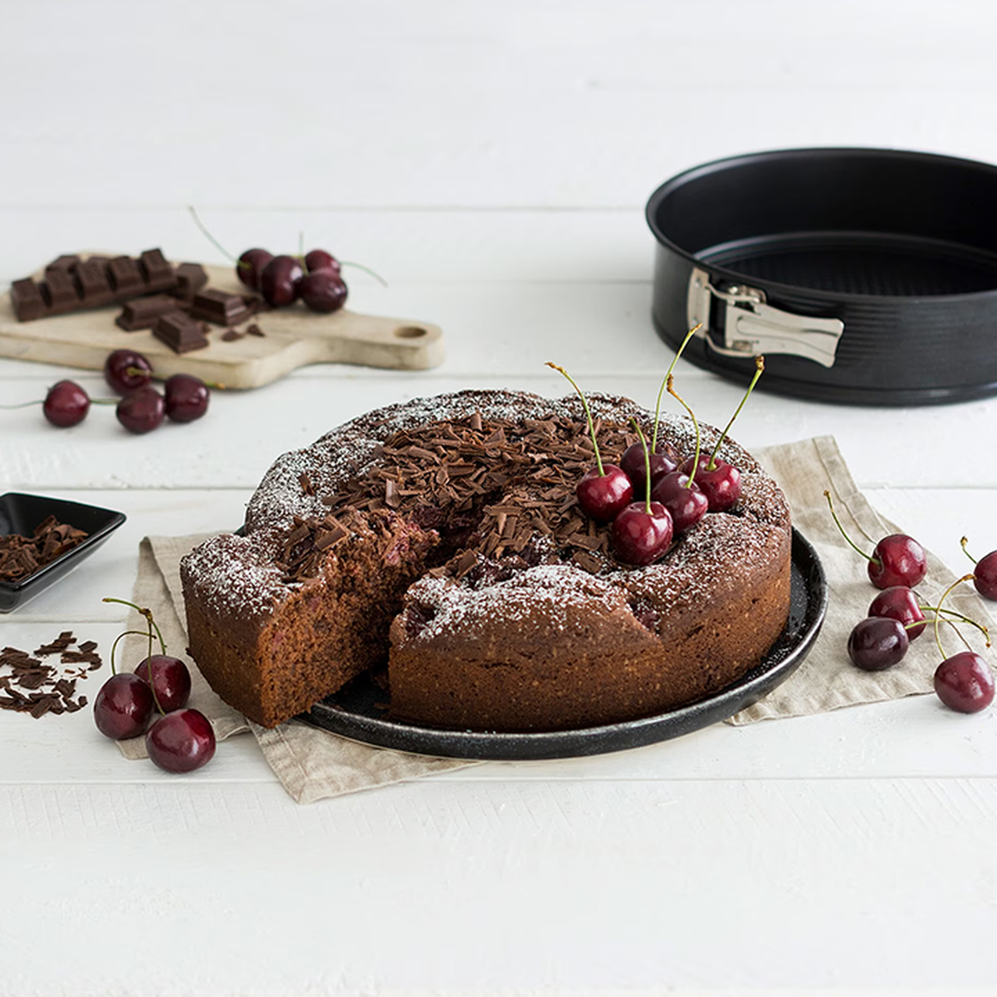 Schokoladenkuchen mit Kirschen und Schokospänen als Belag, teilweise aufgeschnitten, auf einem schwarzen Teller. Ringsherum sind Kirschen und Schokoladenstücke verstreut.