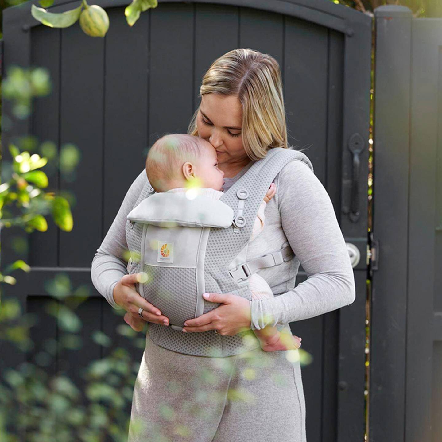 Die Mutter hält ihr Baby in einer grauen Babytrage im Arm. Sie steht draußen in der Nähe eines Tores, umgeben von Grün. Beide tragen hellgraue Kleidung.