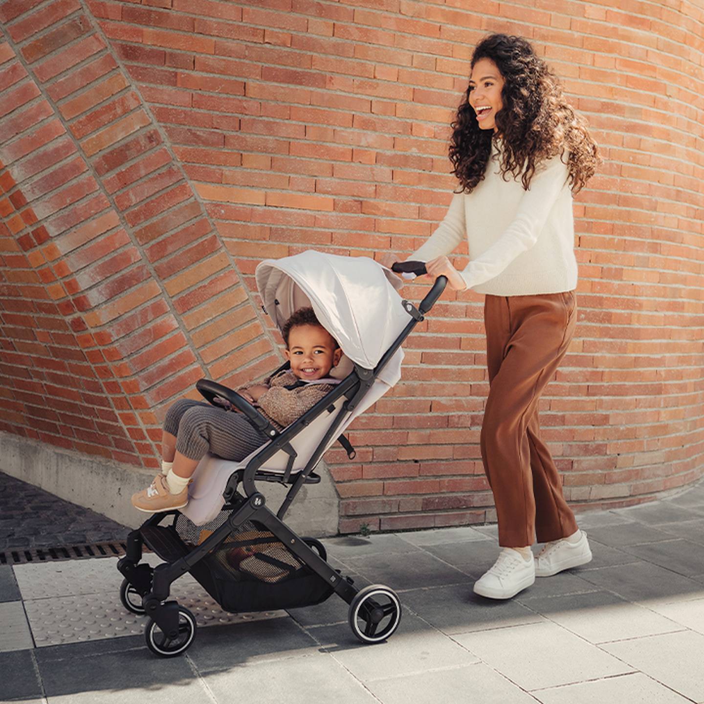 Eine lächelnde Frau schiebt einen Kinderwagen an einer Backsteinmauer entlang. Das Kind lächelt ebenfalls und wirkt entspannt.