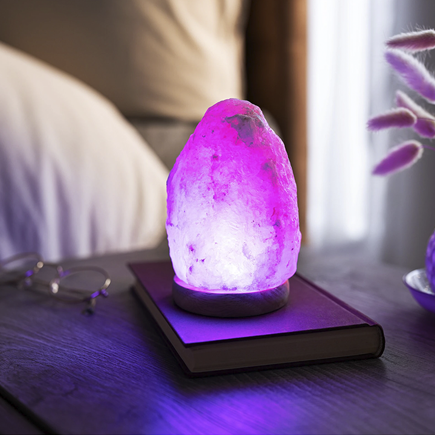 Eine leuchtend rosa Salzlampe steht auf einem geschlossenen Buch auf einem Nachttisch aus Holz, neben einer Brille und dekorativem Gras.