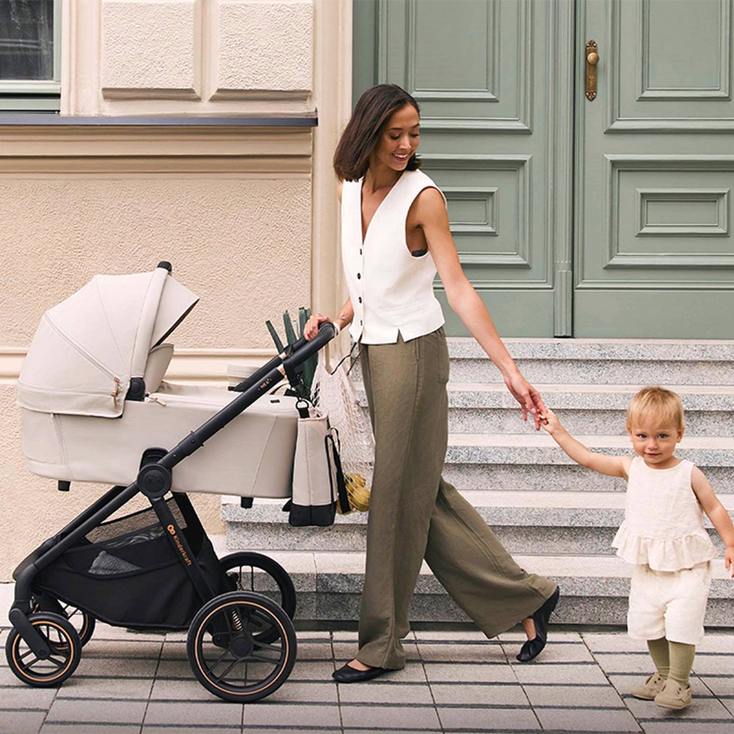 Eine Frau in weißer Weste und grüner Hose geht mit einem Kleinkind und einem Kinderwagen auf einem Bürgersteig in der Stadt und lächelt vor einer grünen Tür.