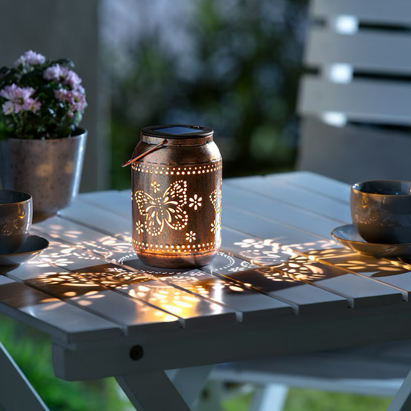 Eine Laterne mit Schmetterlingsmotiven wirft kunstvolle Schatten auf einen weißen Gartentisch, neben dem eine Topfpflanze und zwei Tassen stehen.