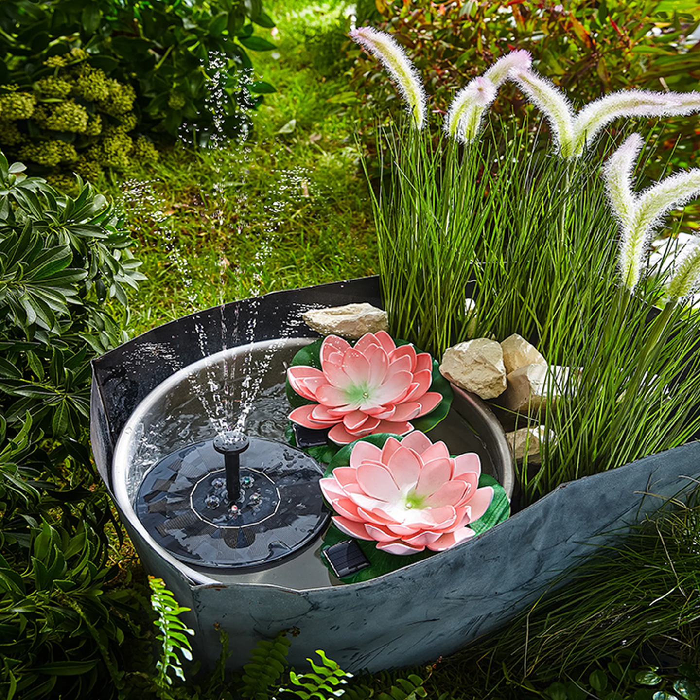 Ein kleiner Gartenbrunnen mit zwei rosafarbenen Lotusblüten und Wasserstrahlen, umgeben von Gräsern und Steinen.