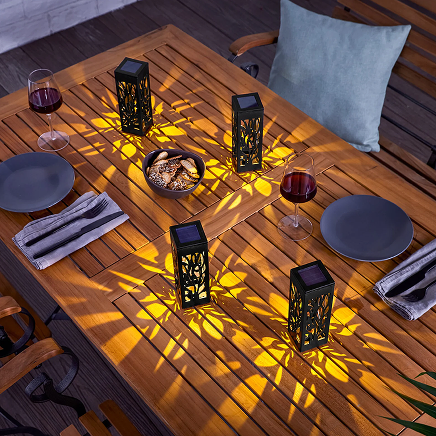 Ein Holztisch, gedeckt mit kunstvoll gemusterten Laternen, die Schatten in Blattform werfen, zwei Gläsern Rotwein, Tellern, Servietten und einer Schale mit Keksen.