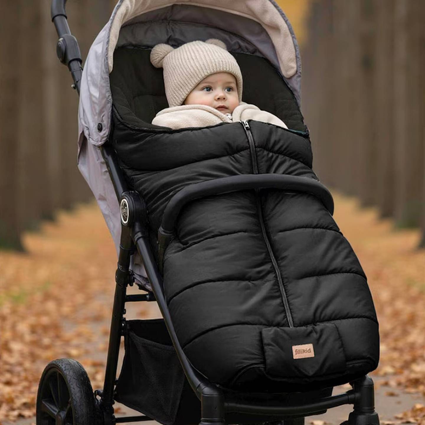 Ein Baby in einem Kinderwagen mit einem Winter-Fußsack, umgeben von einem mit Herbstblättern bedeckten Weg in einer Waldlandschaft.