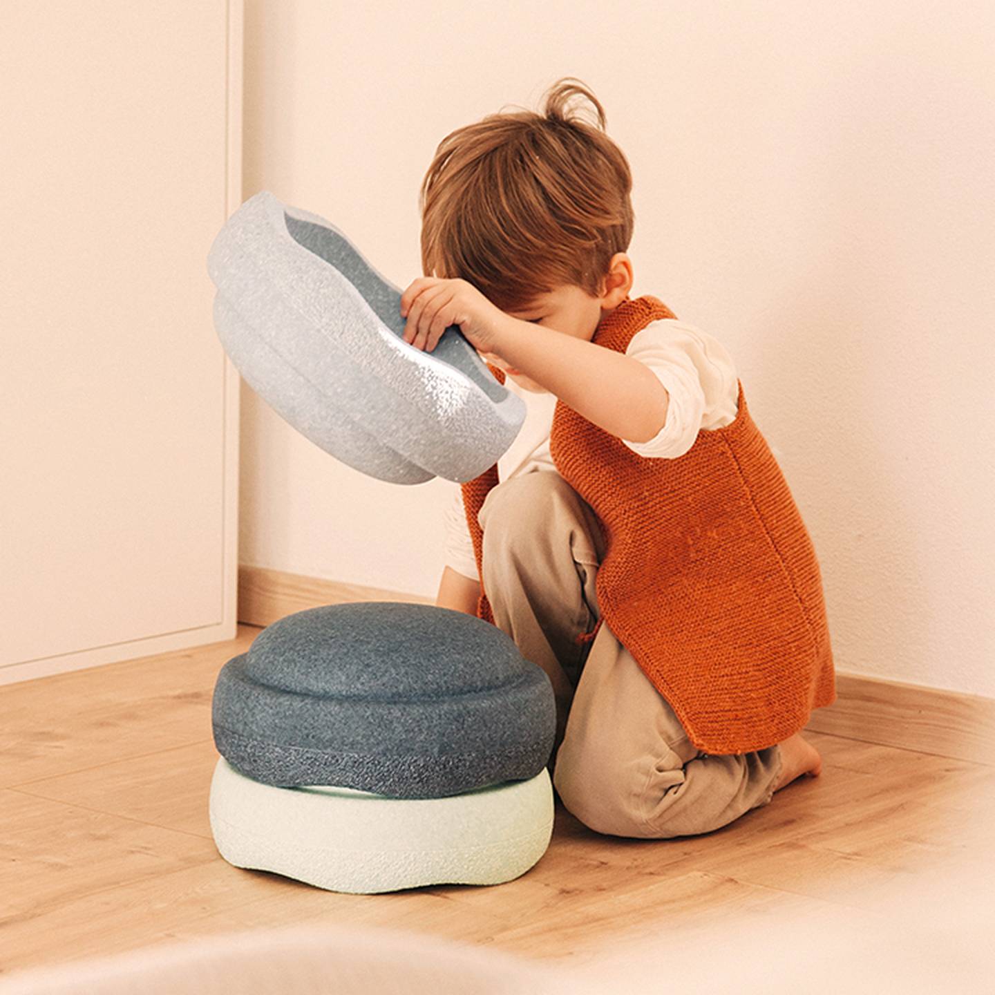 Ein Kind in orangefarbener Weste spielt drinnen auf einem Holzboden mit stapelbaren, kissenartigen Gegenständen.