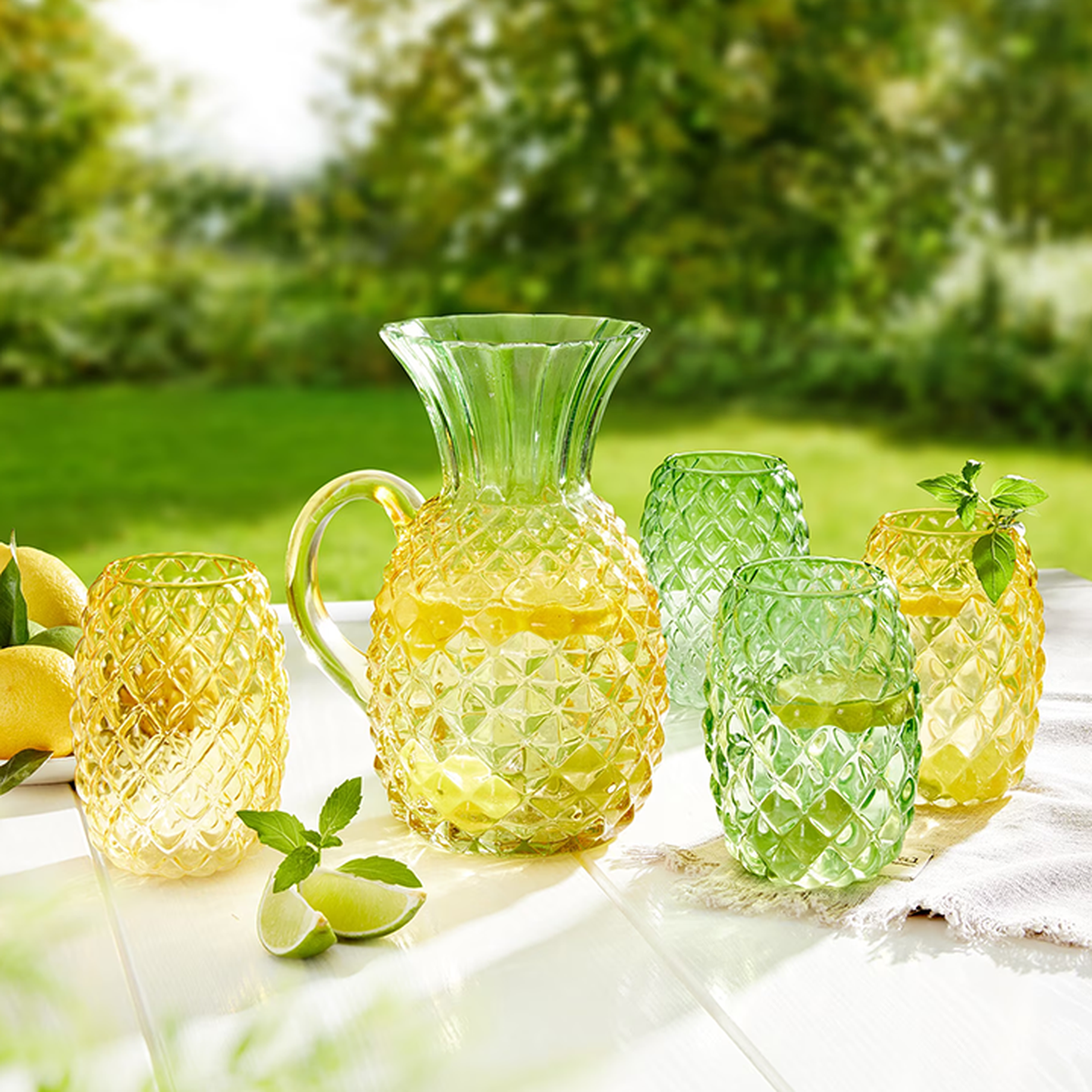 Eine bunte Kanne aus strukturiertem Glas sowie passende Gläser, gefüllt mit Limonade, Zitronen und Minze, auf einem Tisch im Freien vor einem verschwommenen grünen Hintergrund.