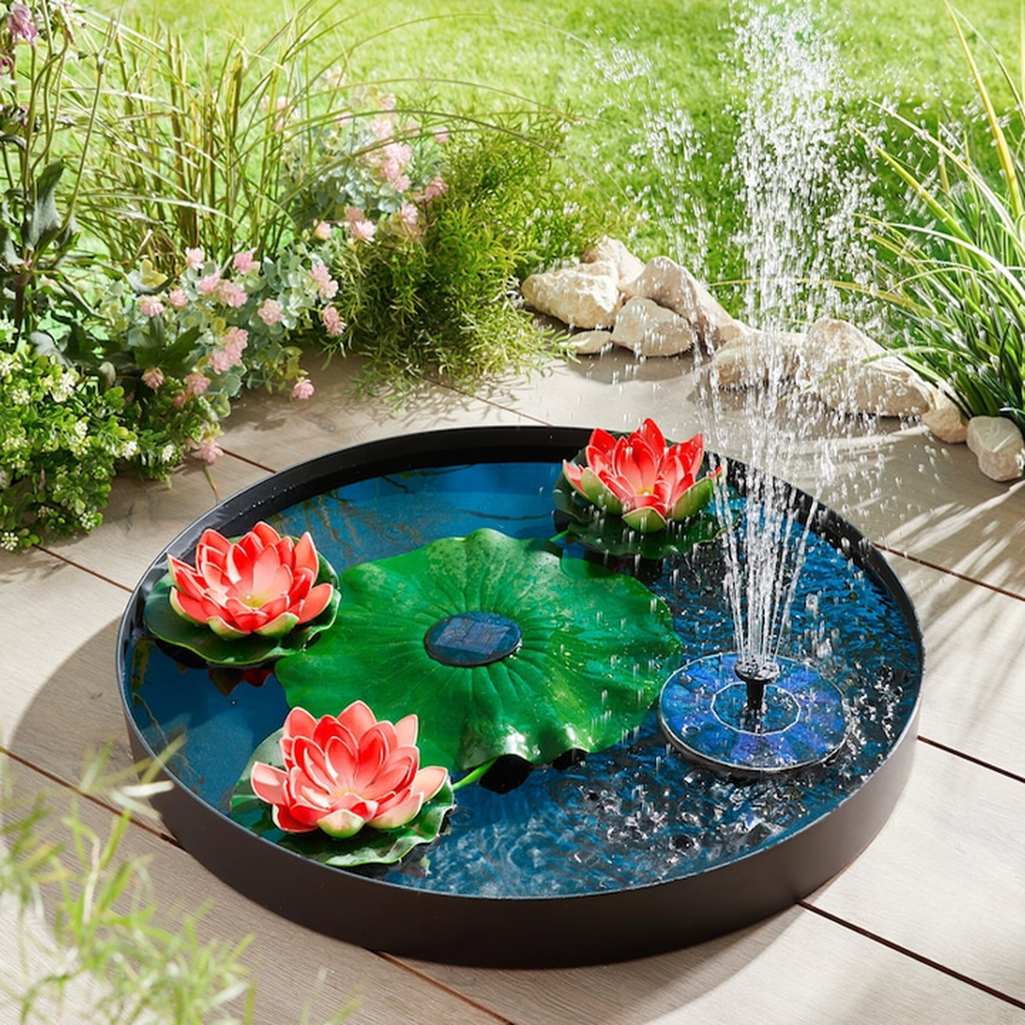 Ein runder Springbrunnen mit schwimmenden roten Lotusblumen, grünen Seerosenblättern und einem solarbetriebenen Wasserstrahl, umgeben von üppigen Pflanzen und Steinen.