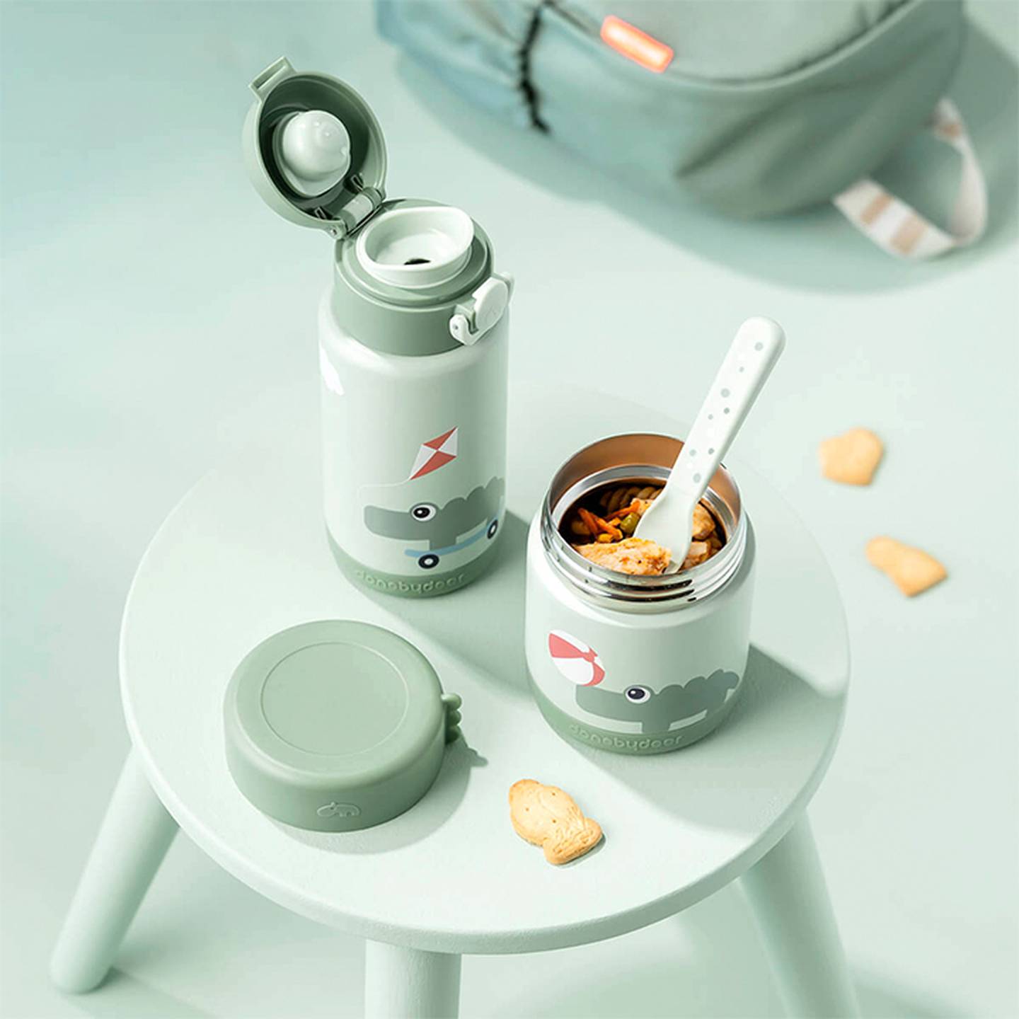 Zwei niedliche Thermoskannen mit Tiermotiven stehen auf einem pastellgrünen Hocker und sind mit Nudeln gefüllt. Daneben liegen ein Löffel und Kekse in Tierform.