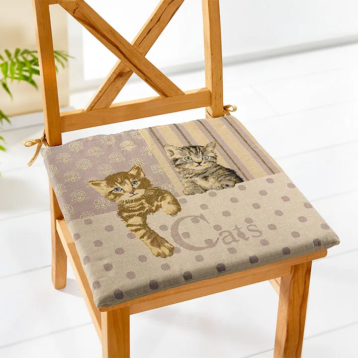 Stuhl mit einem Kissen mit zwei Kätzchenillustrationen und dem Wort „Cats“ vor einem hellen Boden mit einer Pflanze im Hintergrund.