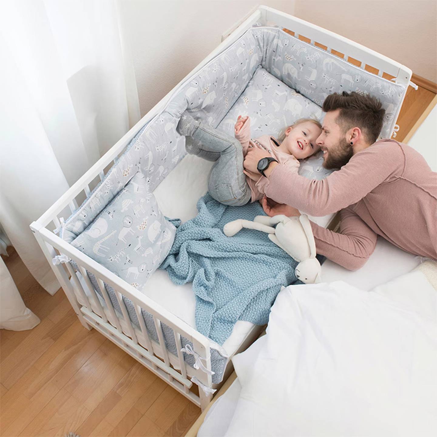 Ein Vater kuschelt mit seinem Kind in einem Kinderbett mit Kissen mit Tiermuster und einer blauen Decke, neben einem größeren Bett.