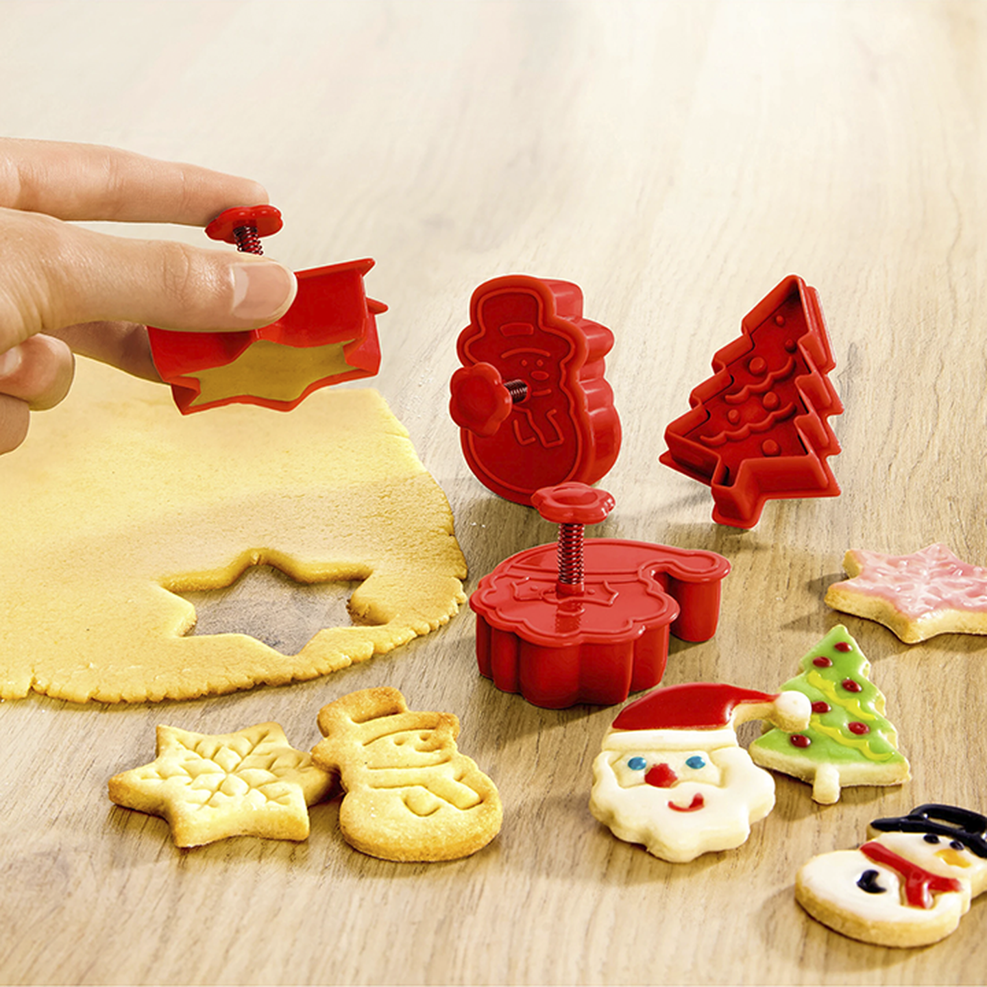 Hand, die rote Ausstechformen auf Teig mit festlichen Keksen in Form von Sternen, Schneemännern, Bäumen und Weihnachtsmännern auf einer Holzoberfläche verwendet.