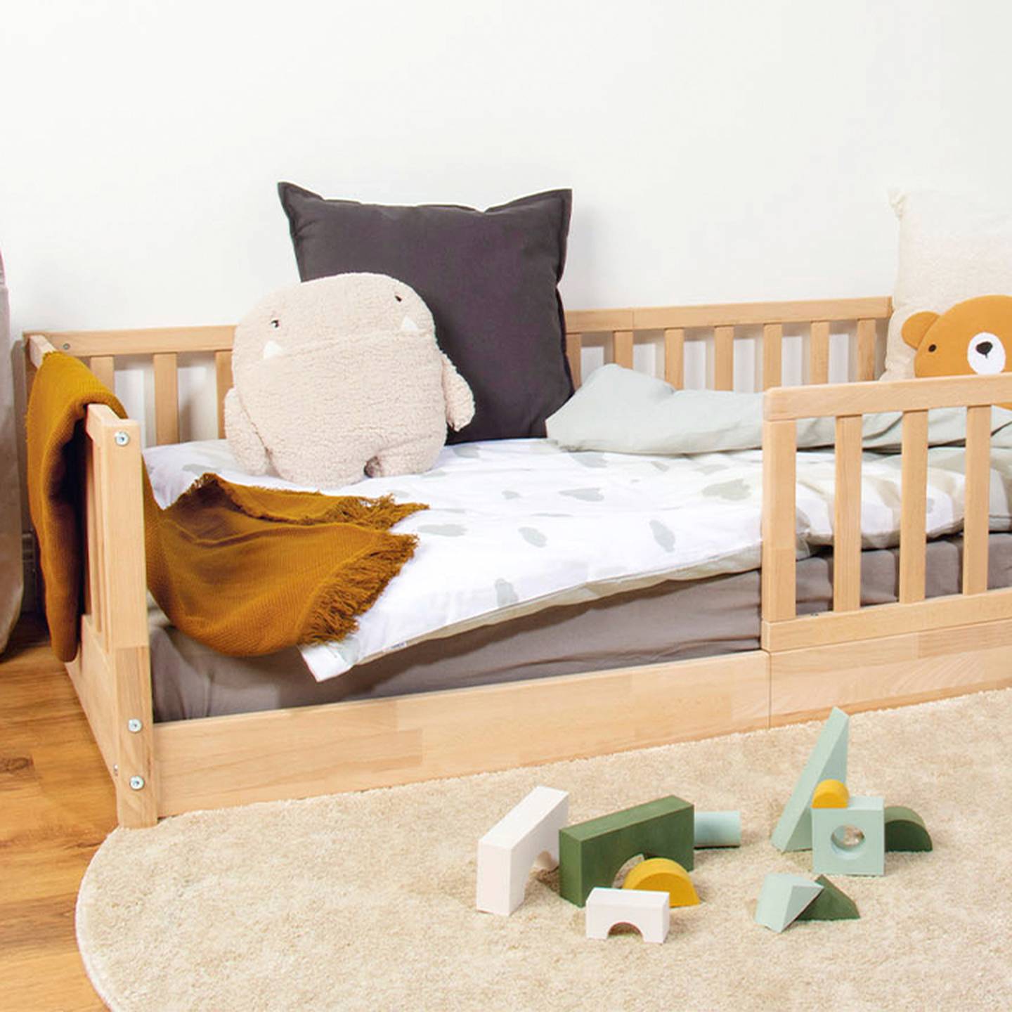 Ein hölzernes Kleinkinderbett mit Kissen und einem weichen Plüschtier, darunter eine senffarbene Decke. Auf einem runden beigen Teppich liegen verstreut Holzspielzeuge.